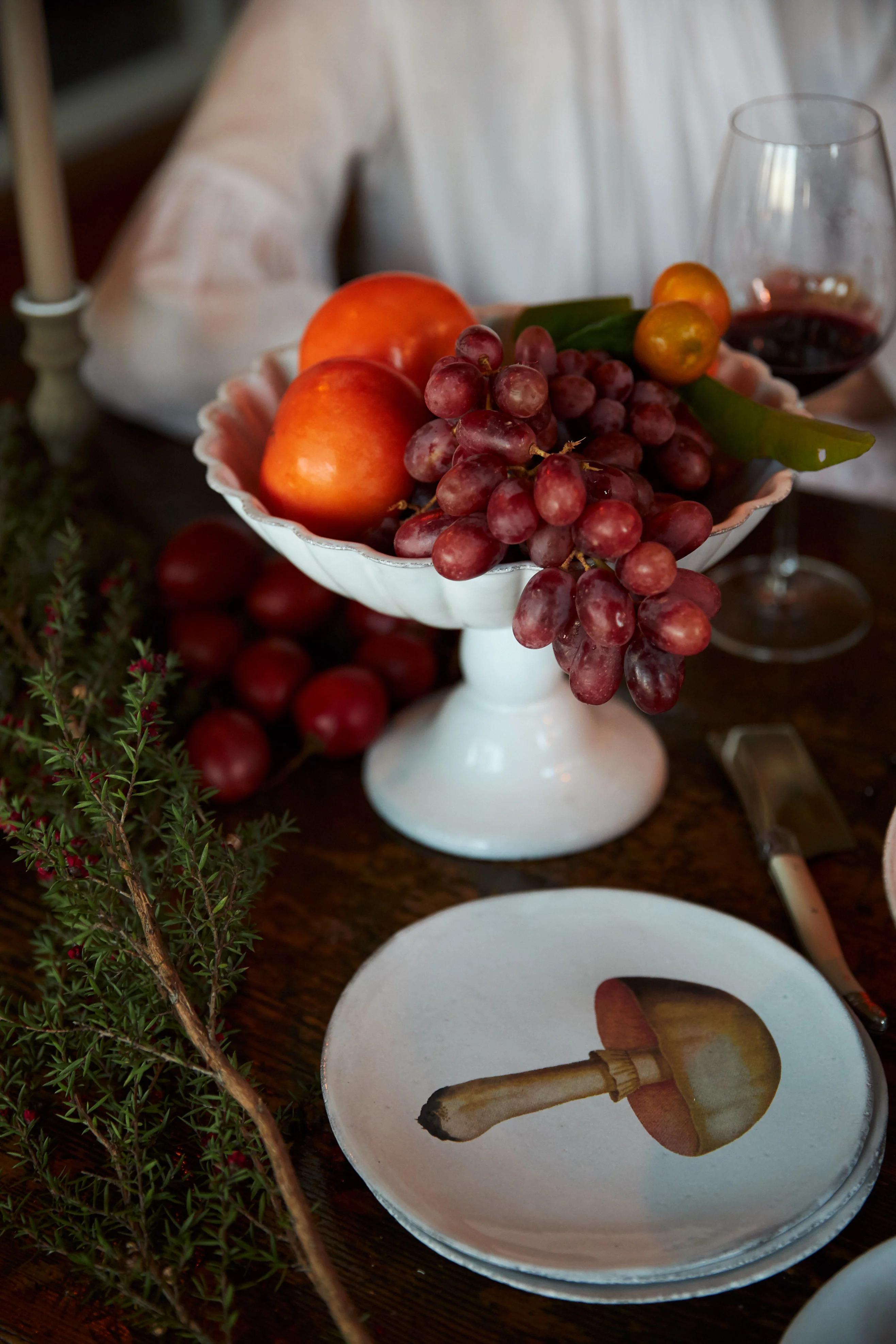 White ceramic bowl with oranges, grapes, and lime on wooden table. Wine glass, greens, and mushroom-illustrated plate nearby. Person in white shirt in background, possibly preparing Fruit Tarte Tatin.