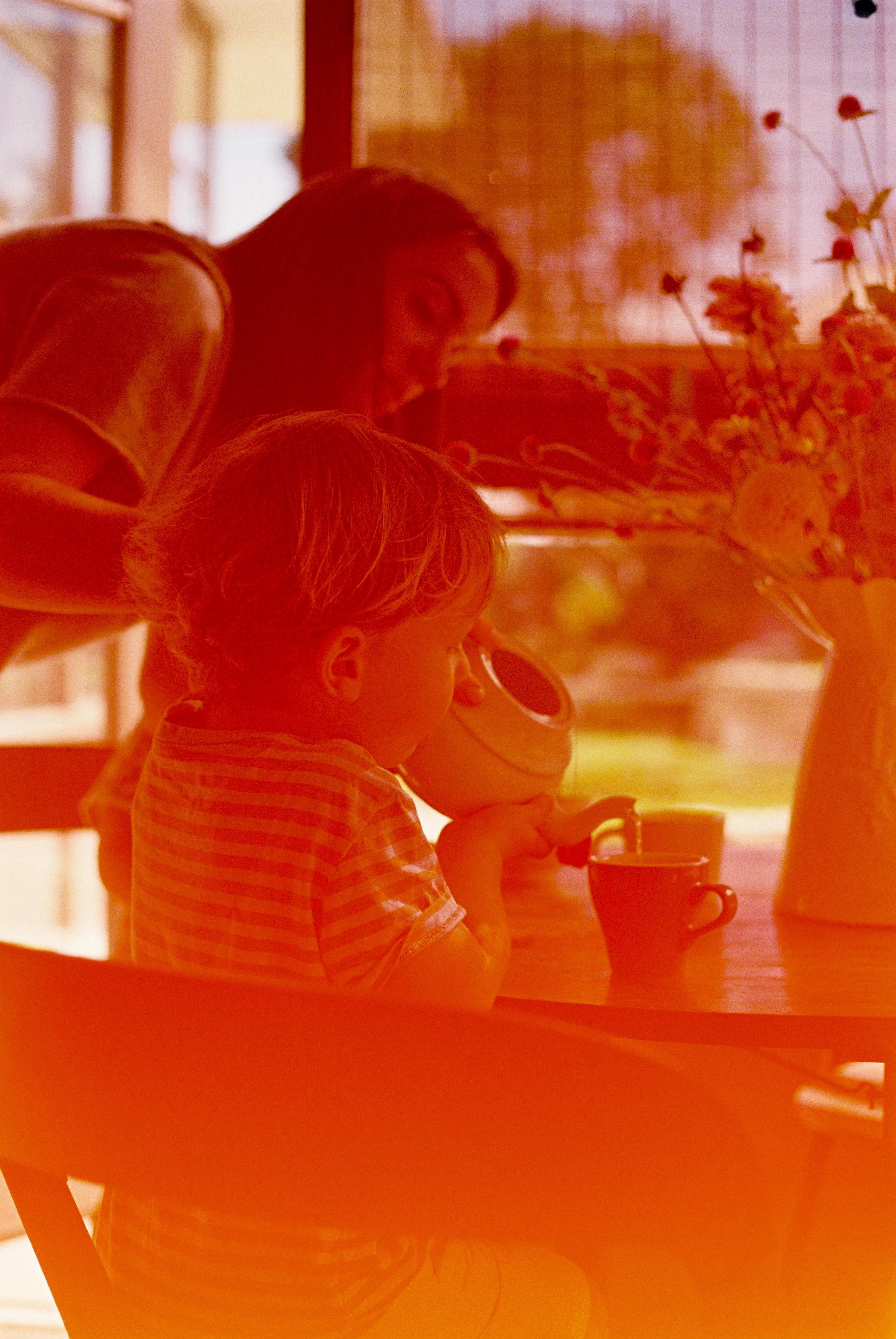 Young child drinks from small cup. Olivia Moon leans over, smiling. Sunlit scene with flower vase and window in background.