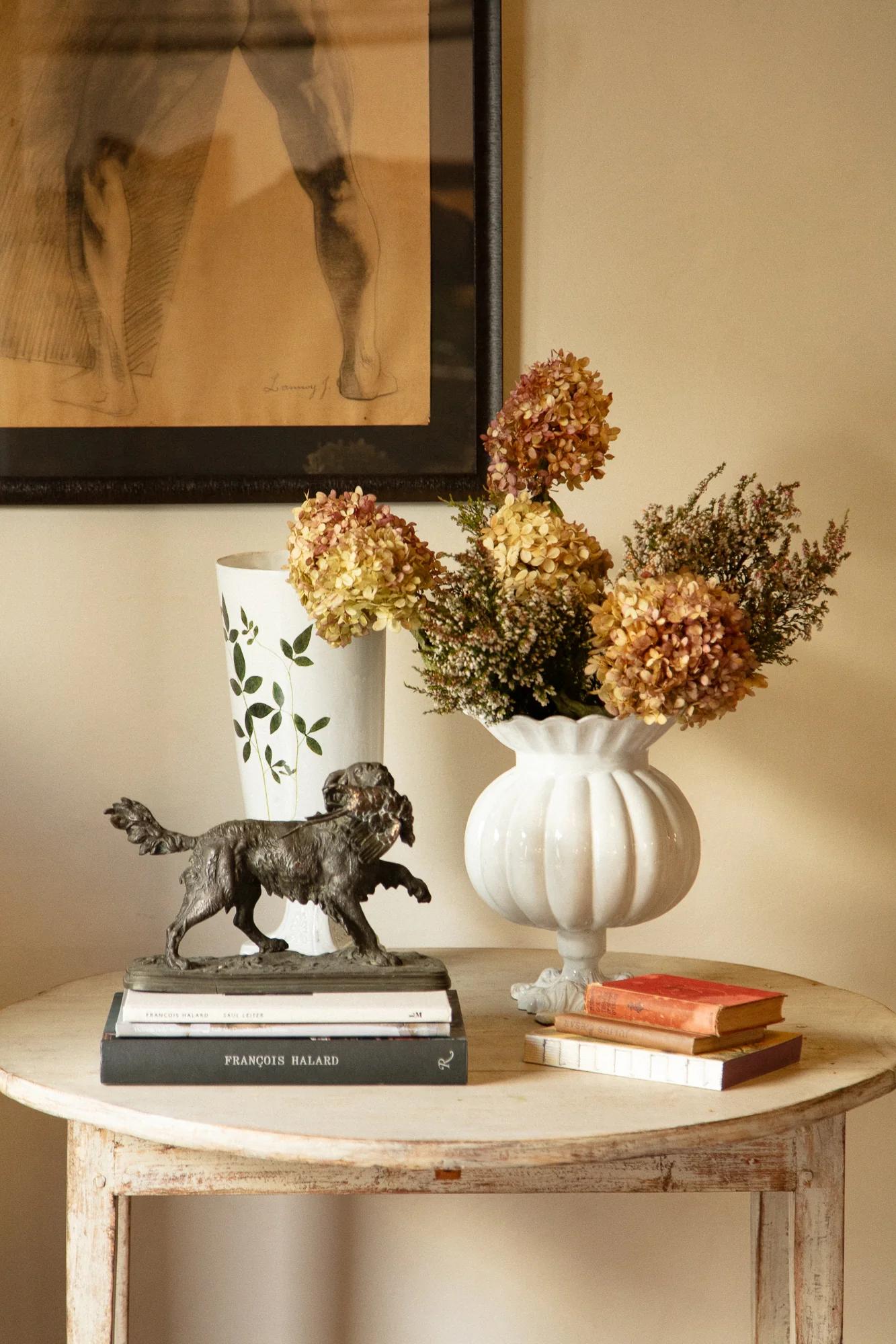 Wooden table with metal dog figurine, open book, stack of books, white vase with dried hydrangeas. Tall vase and framed sketch behind.