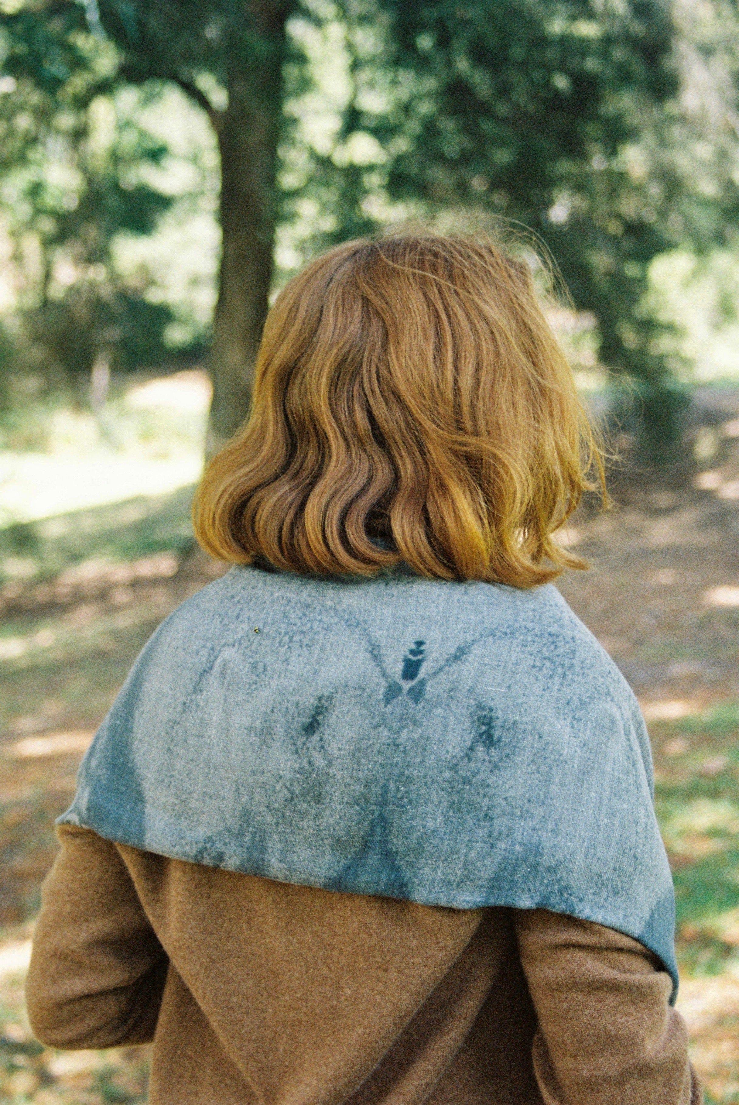 Auburn-haired person in brown coat and blue shawl, back to camera. Standing in sunlit New Zealand forest.