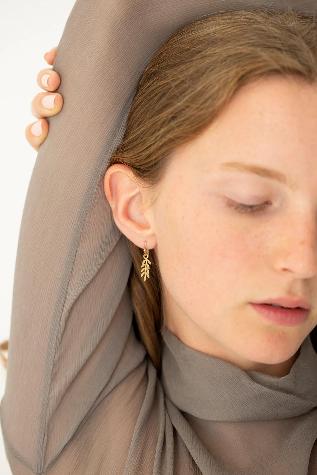 Person in sheer grey fabric, arm raised showing Charlotte Penman gold leaf earring. Eyes closed, calm mood. Contemplative atmosphere.