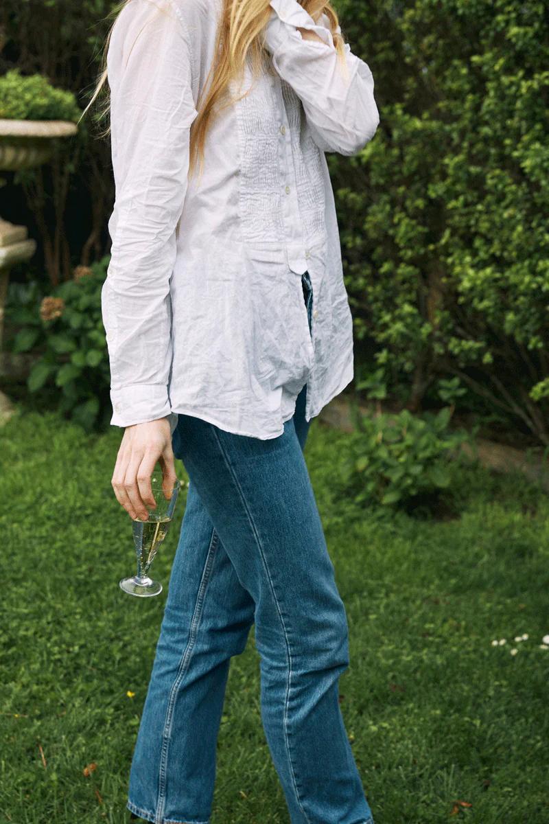 Person in white shirt and jeans holds champagne glass in garden. Lush greenery background suggests outdoor holiday setting.