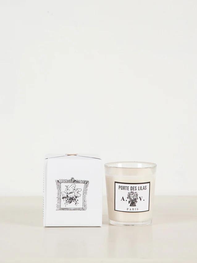 "Porte Des Lilas" white glass candle next to matching box with floral design. Displayed on light surface against white background.