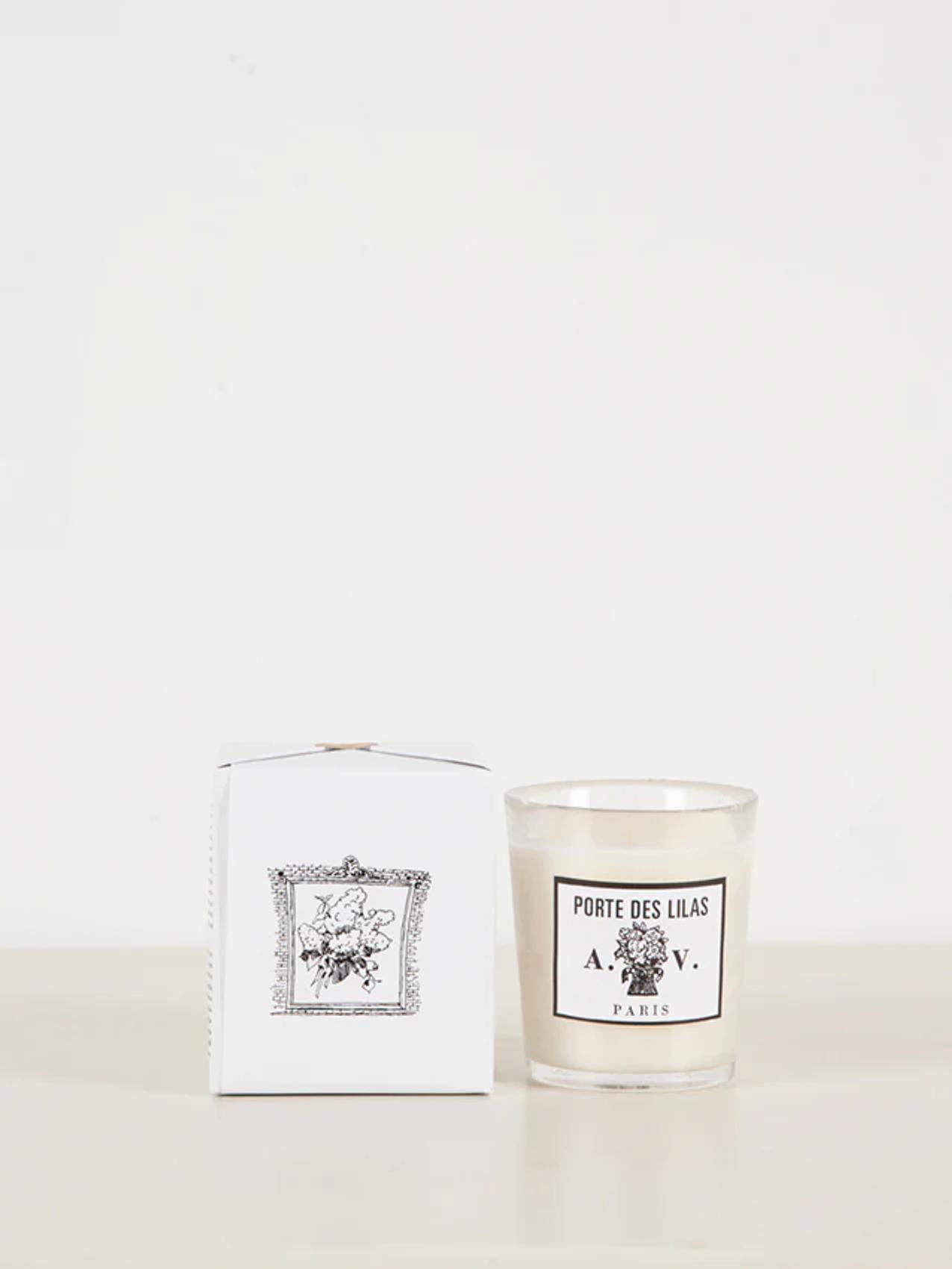 "Porte Des Lilas" white glass candle next to matching box with floral design. Displayed on light surface against white background.