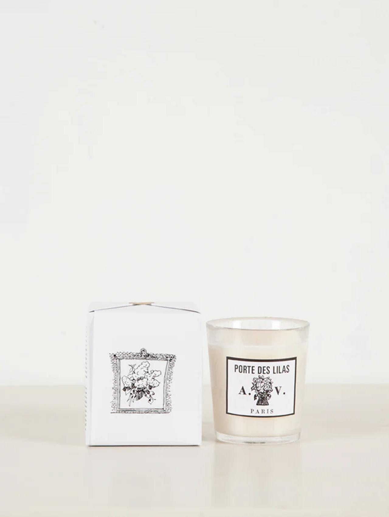 "Porte Des Lilas" white glass candle next to matching box with floral design. Displayed on light surface against white background.