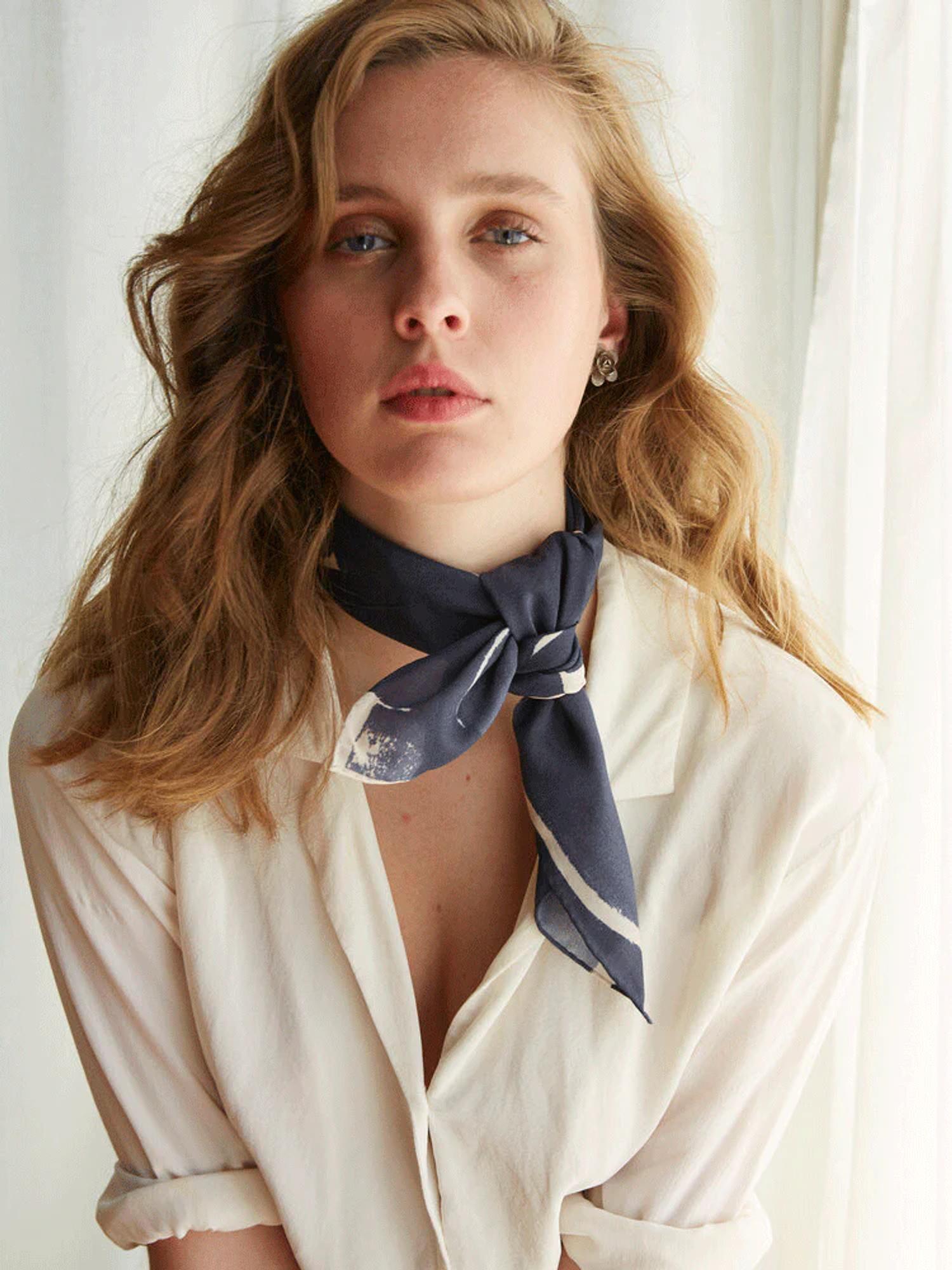Blonde person in cream shirt and patterned Walker & Bing scarf poses in front of light curtain. Neutral expression, looking directly at camera.