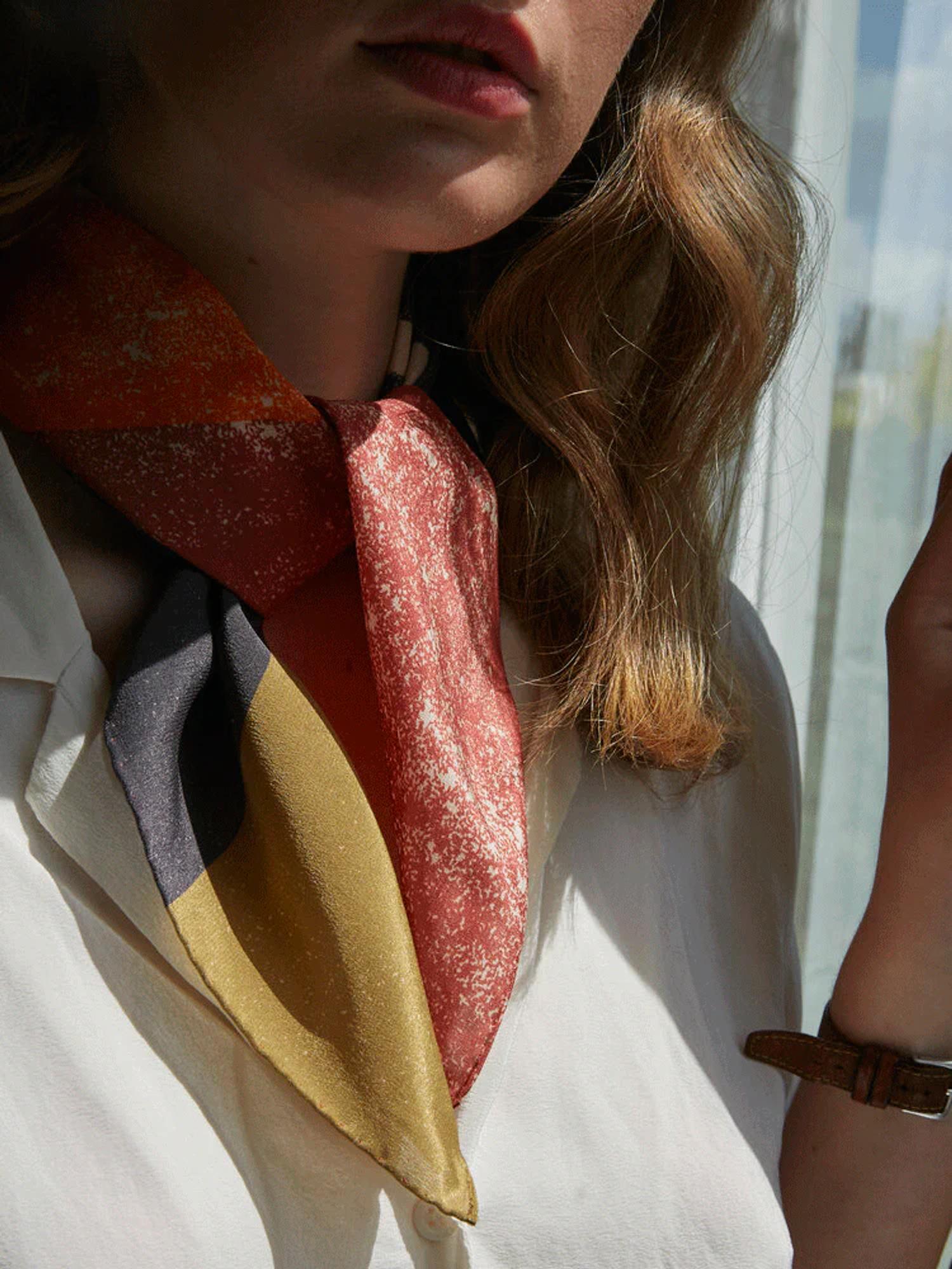 Person in white blouse and vibrant scarf. Light brown wavy hair over shoulder, partially lit by sun. Brown watch visible on raised hand near face.