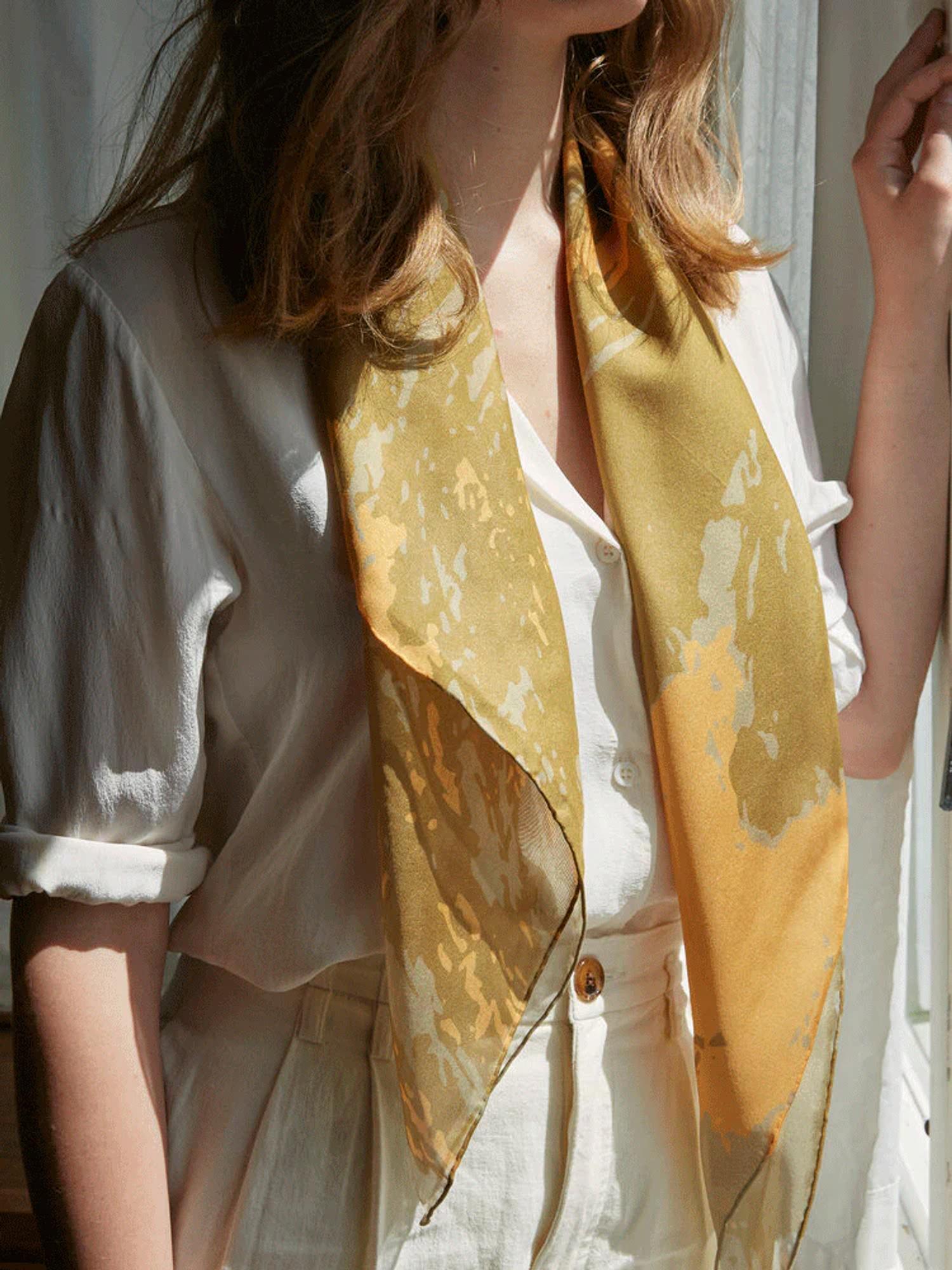 Person in cream outfit wears golden-yellow Walker & Bing scarf with art-inspired pattern. Sunlight casts warm glow, creating serene atmosphere.