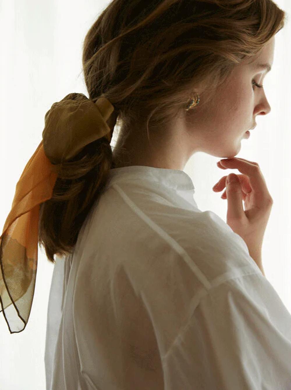 Person with light brown hair in ponytail wears white top and golden Walker & Bing scarf. Touching chin thoughtfully against softly lit background.