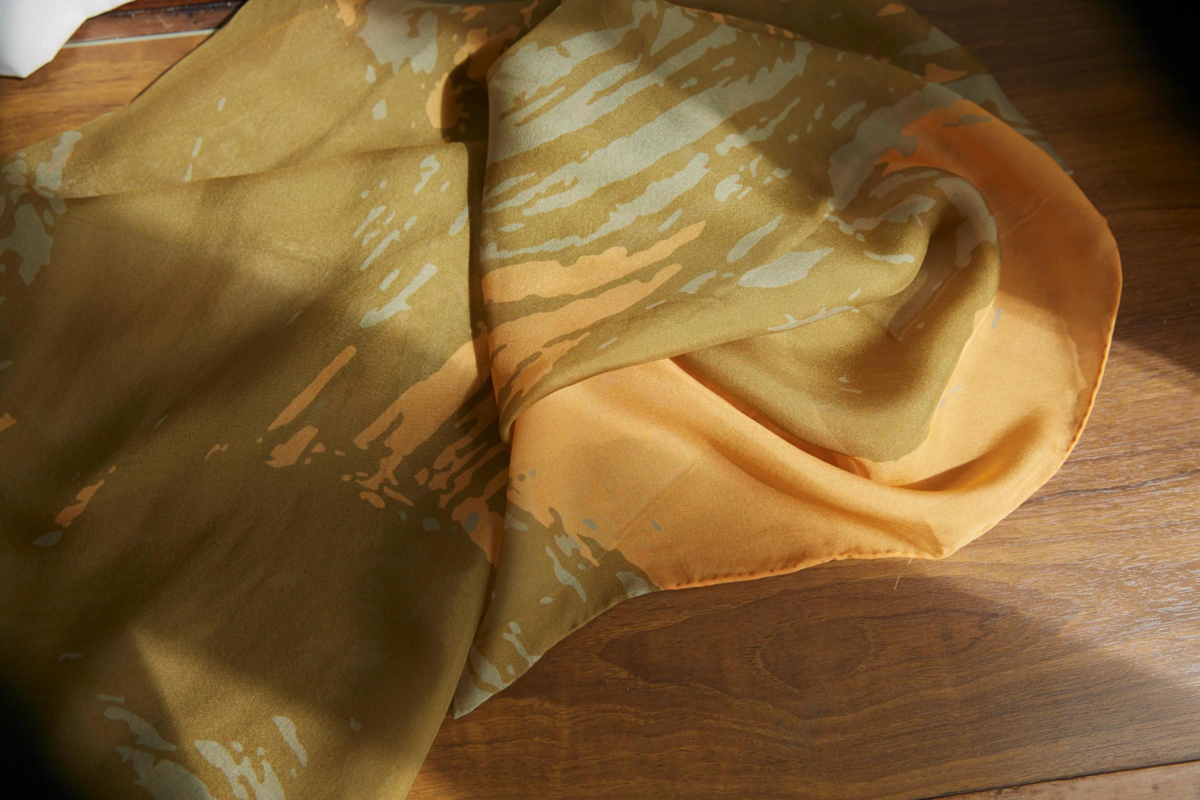 Patterned fabric with olive green, beige, and orange design draped on wood. Sunlight highlights soft folds and rich colours of print-made textile.