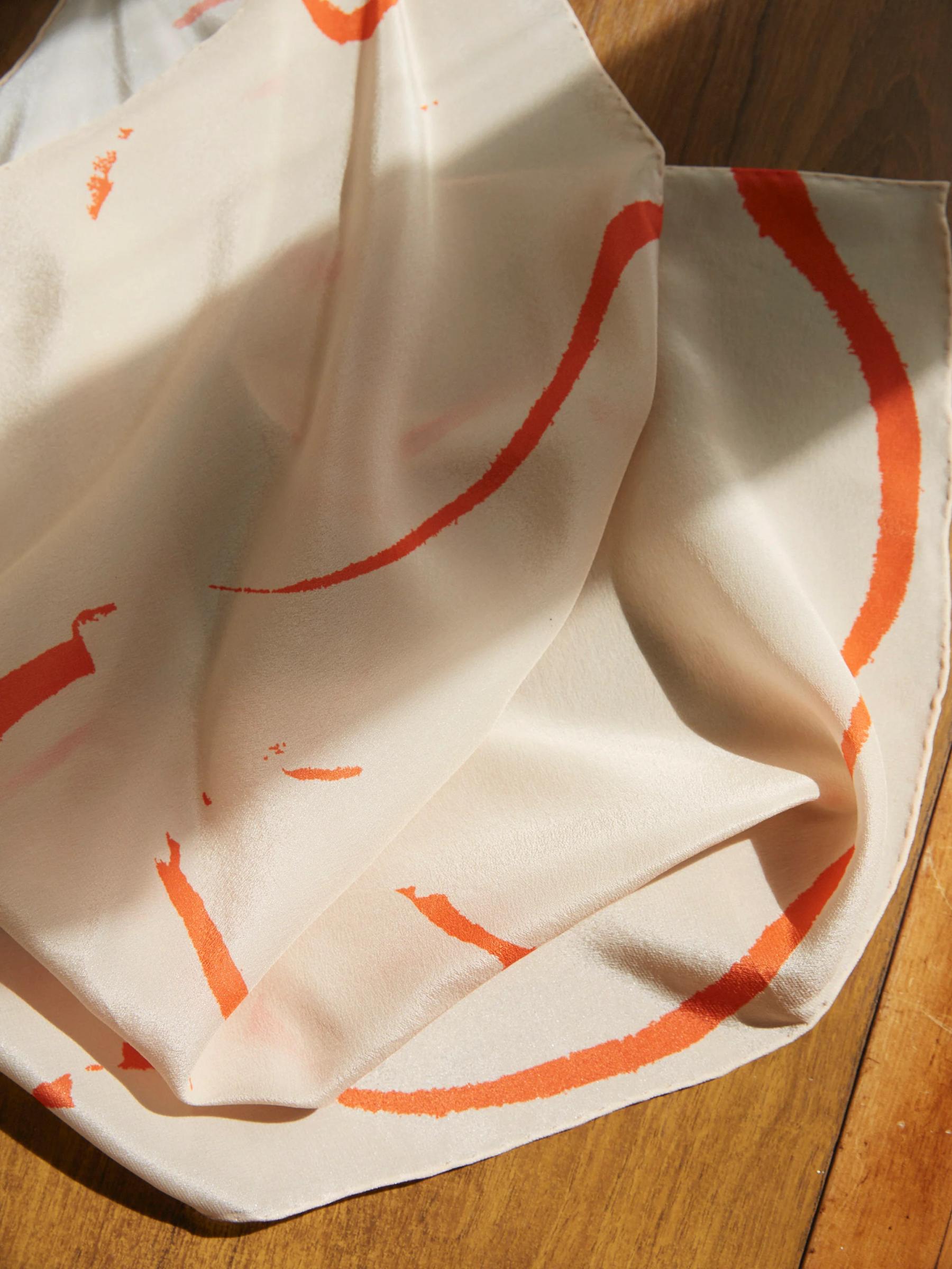 Close-up of white fabric with orange brushstroke patterns. Soft folds on wooden surface create delicate shadows and highlights.