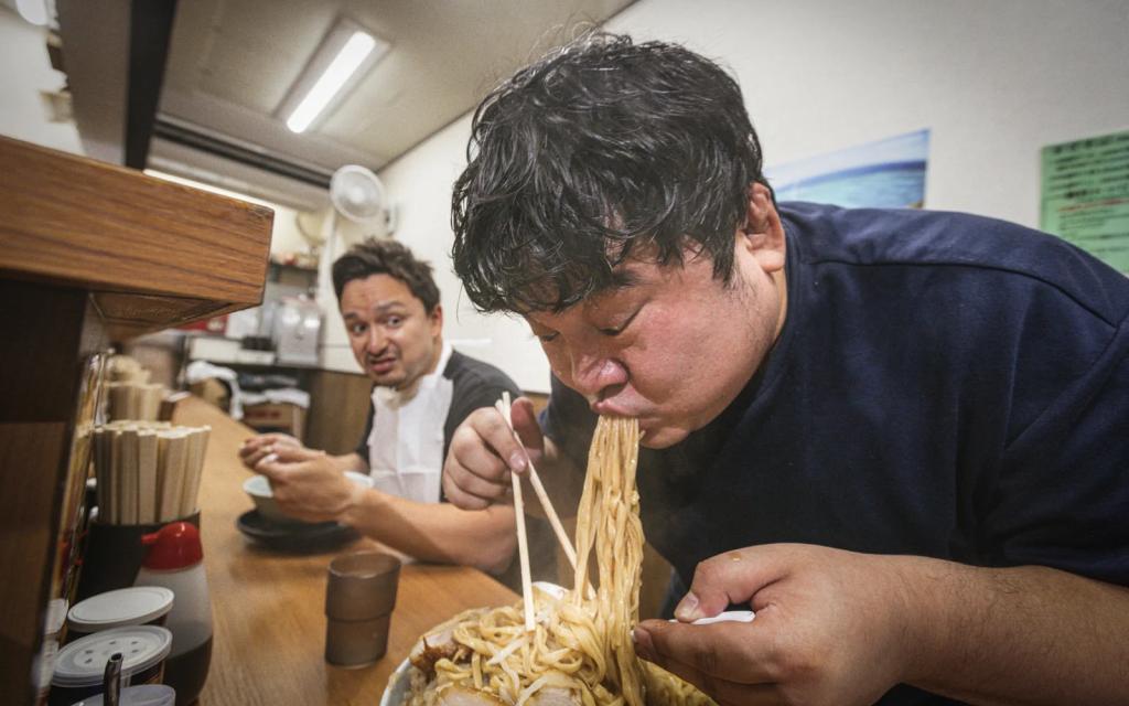 Japanese person eating ramen while another person looks on