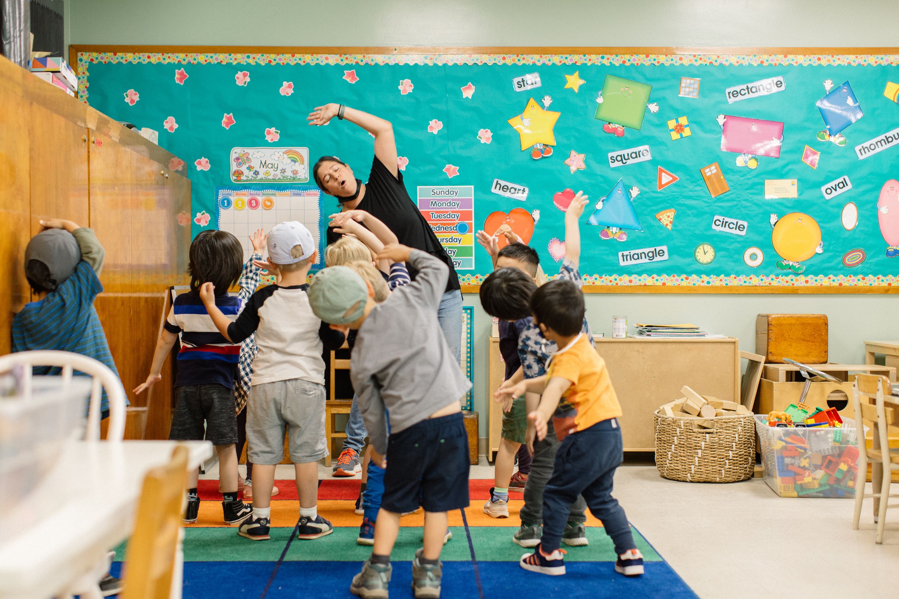 An adult woman stands in a classroom in front of nine children. She is showing them how to stretch.