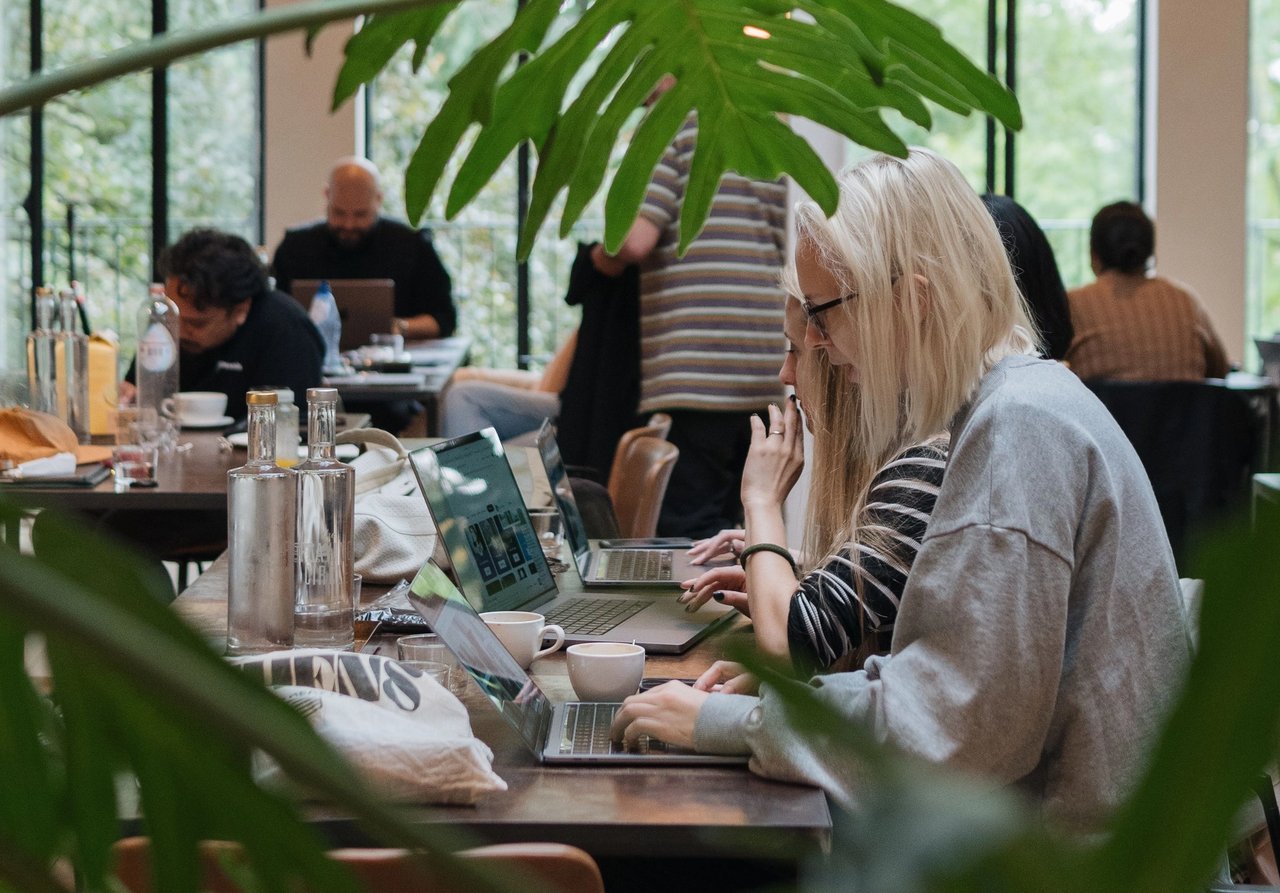 Two colleagues work remotely on laptops from a cafe.