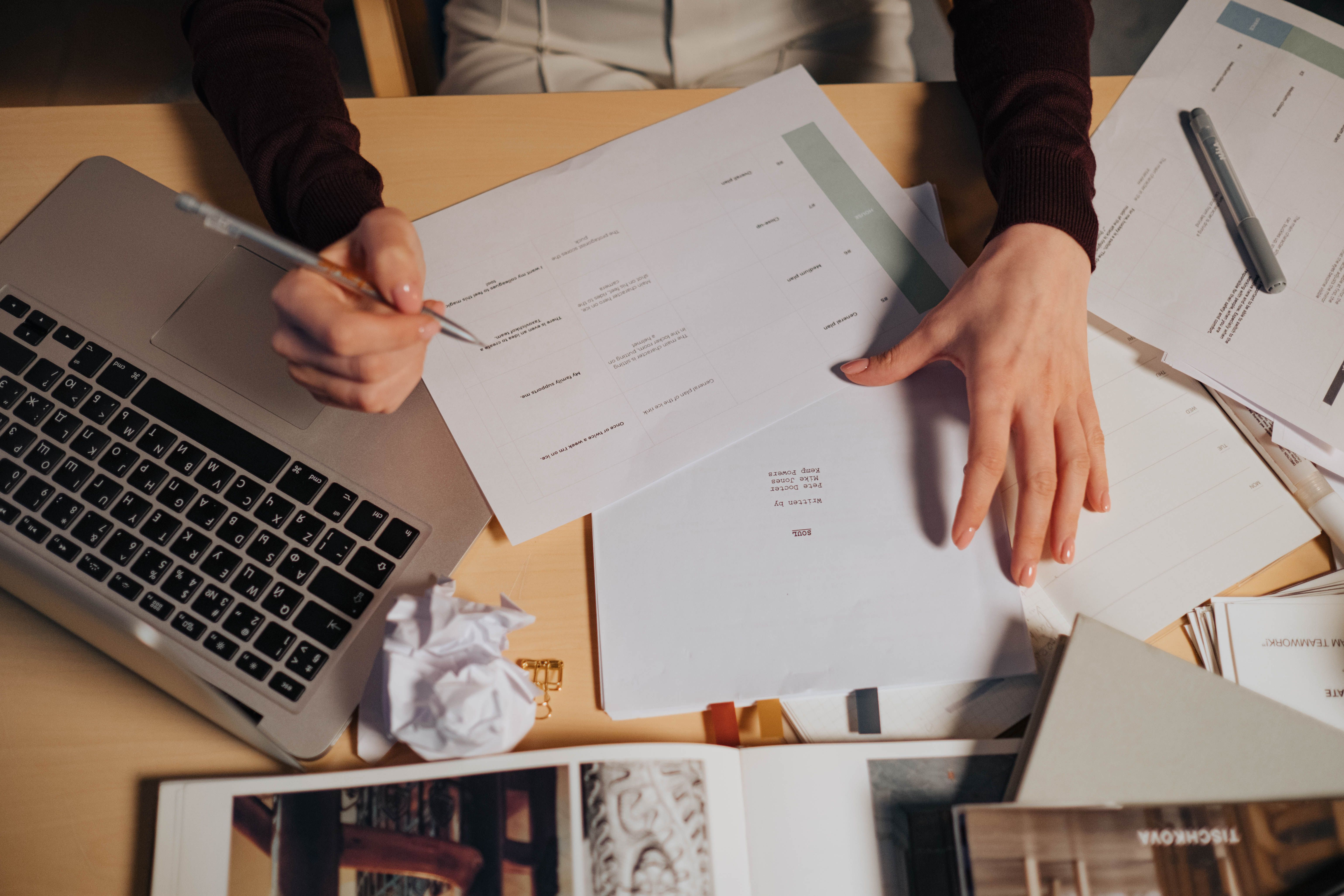 A busy person sat at a desk with their laptop and paper print outs.