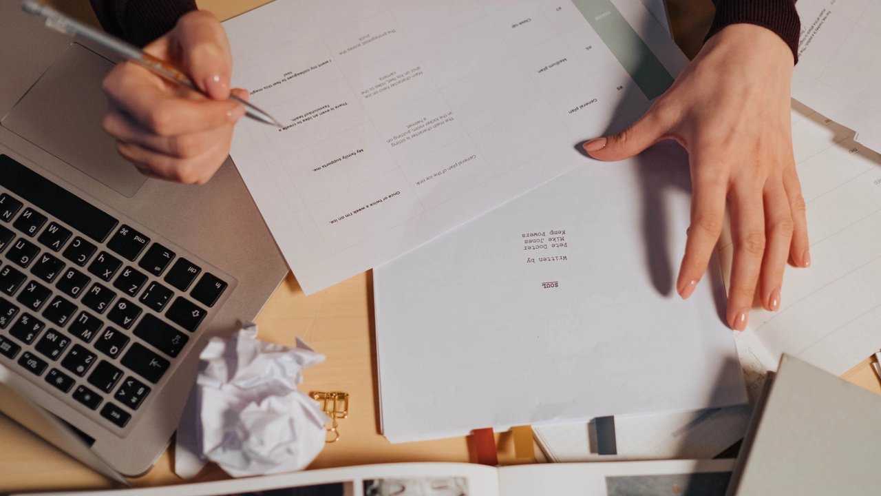 A busy person sat at a desk with their laptop and paper print outs.