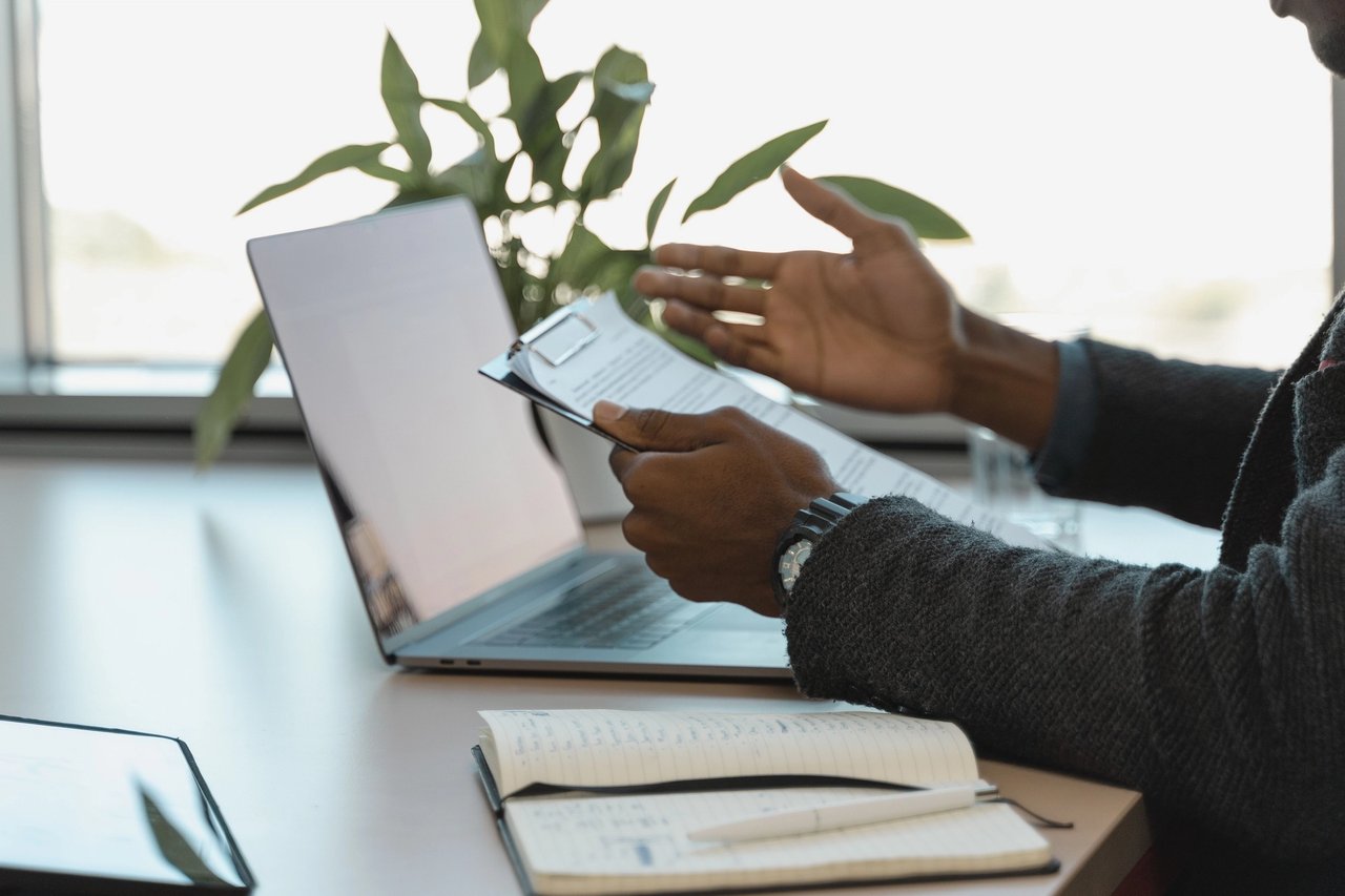 Job seeker sat at laptop confused while studying a printed job description.