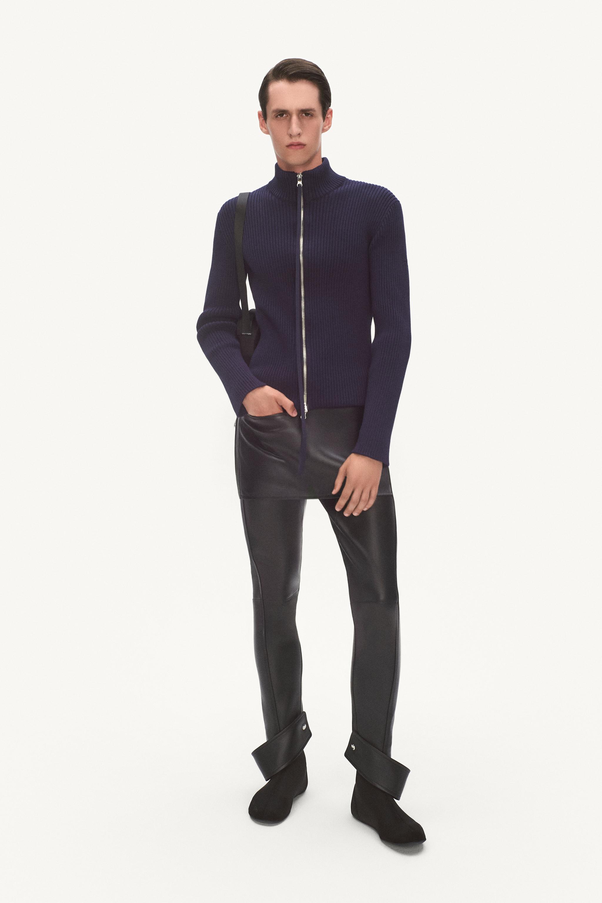 Elegant model wearing a navy blue zip-up sweater and black latex pants on a white background.