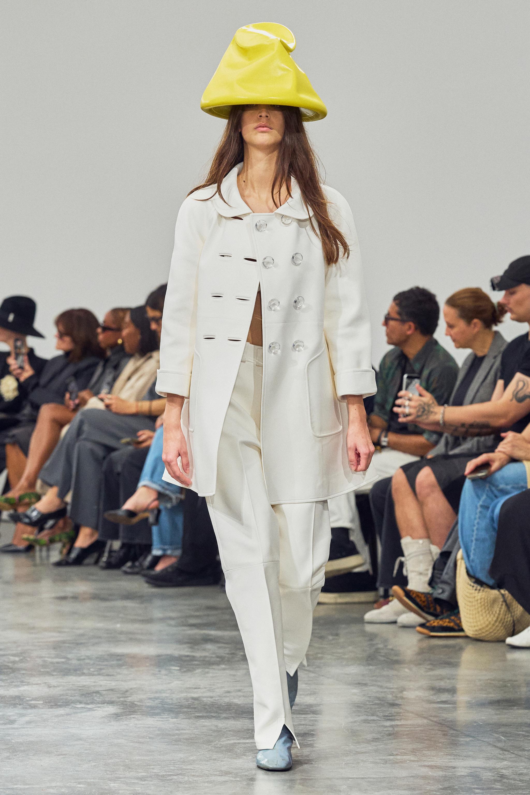 Fashion model wearing a white designer coat and unique yellow hat on runway for Courrèges.