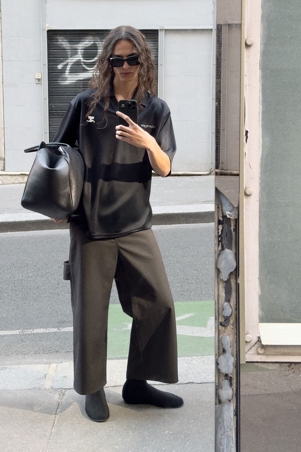 Fashion-forward woman in black Courrèges outfit, carrying a stylish black bag, taking a mirror selfie.
