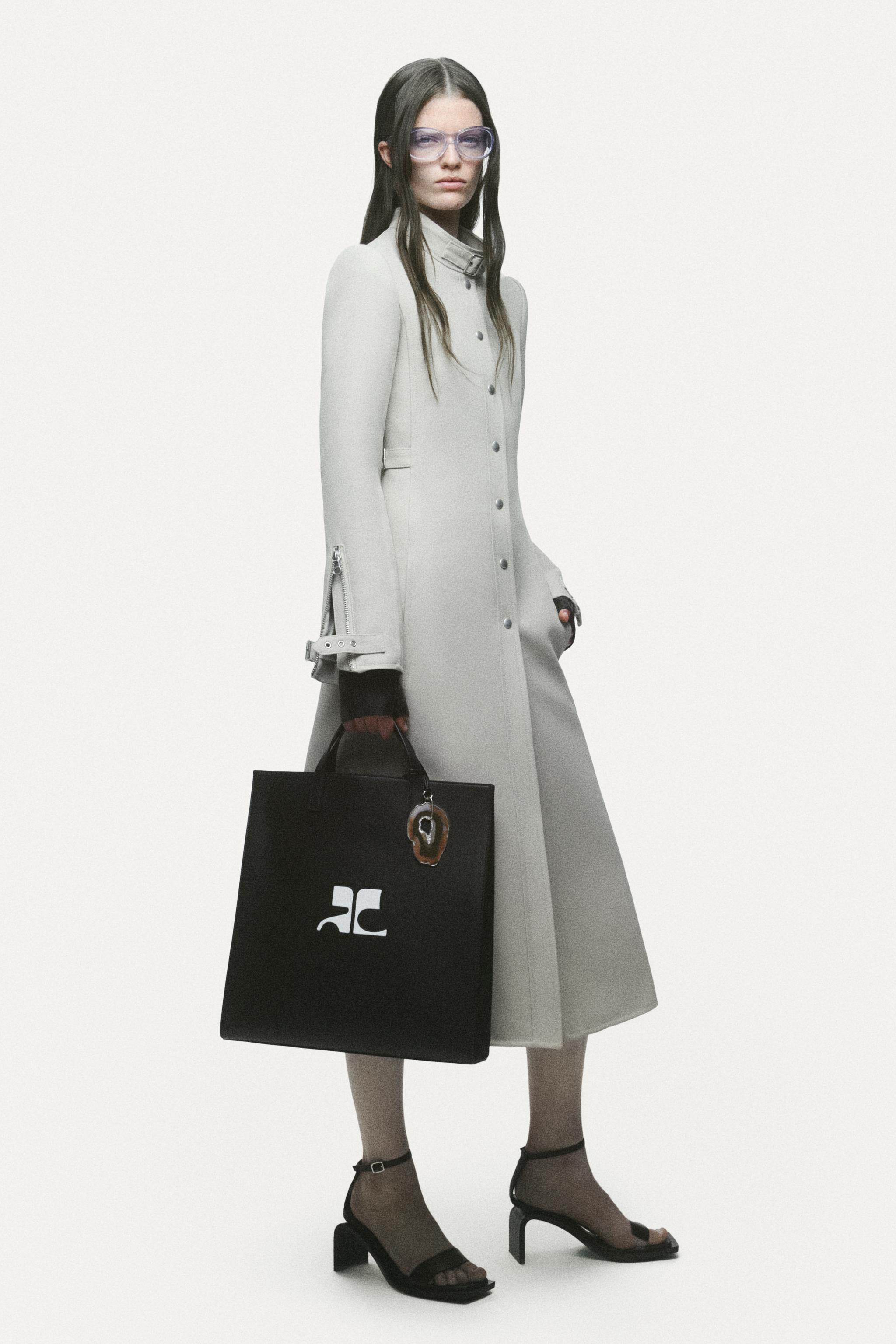 Elegant model in a long coat holding a black Courrèges shopping bag for modern fashion.