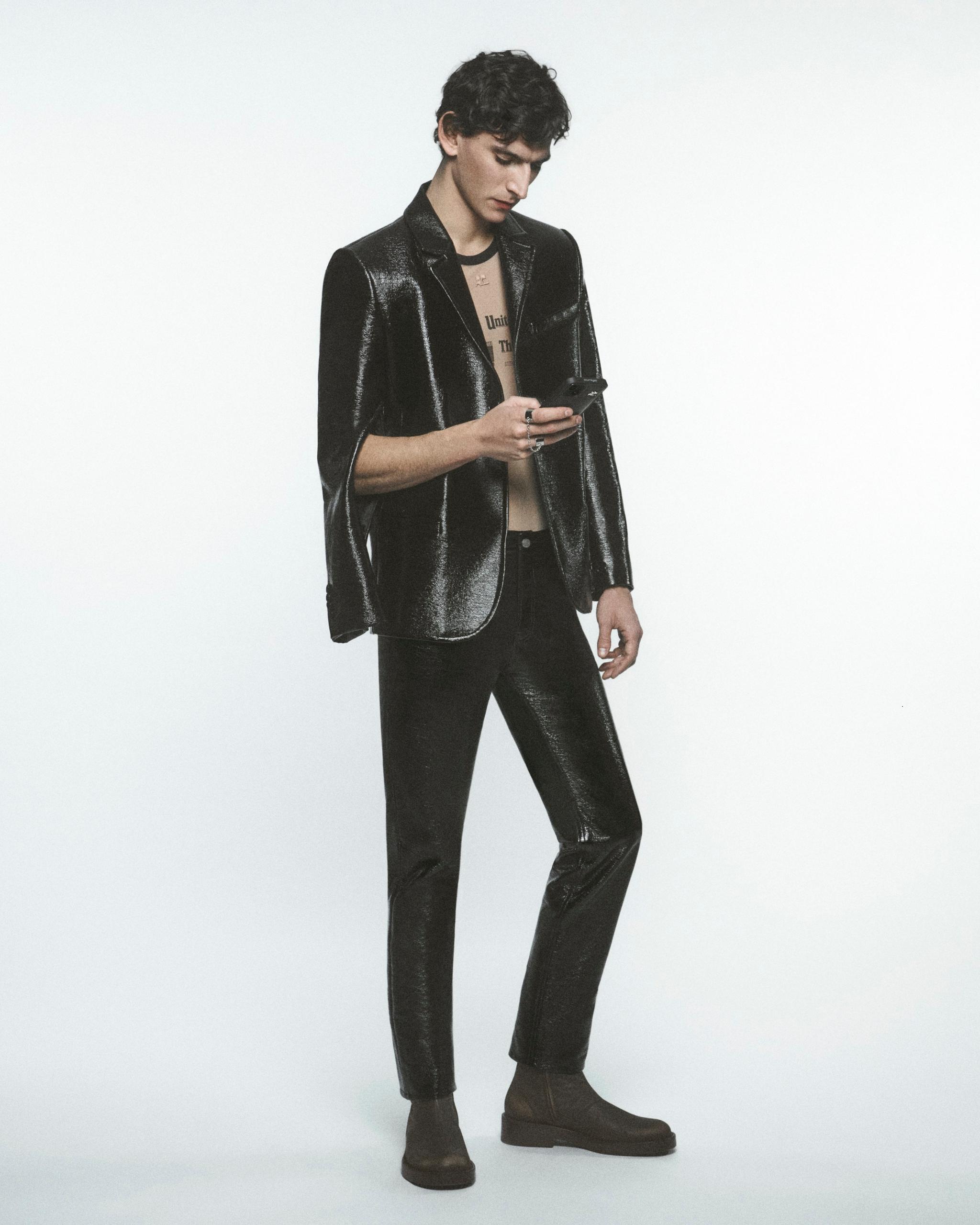 Elegant male model in black shiny Courrèges outfit, using a smartphone, on white background.