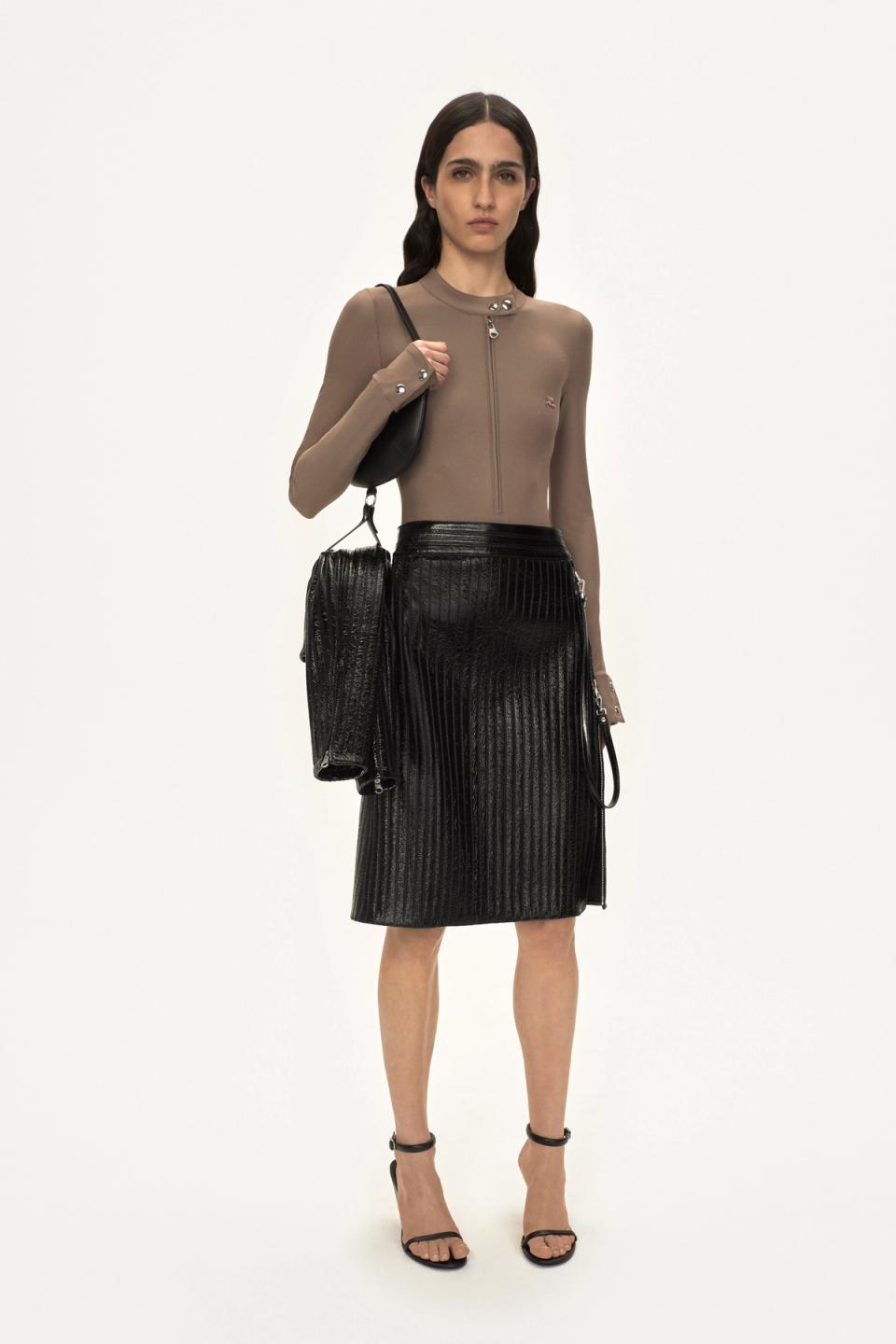 Elegant model in Courrèges fashion brand, wearing a taupe zip-up top with black pleated skirt.