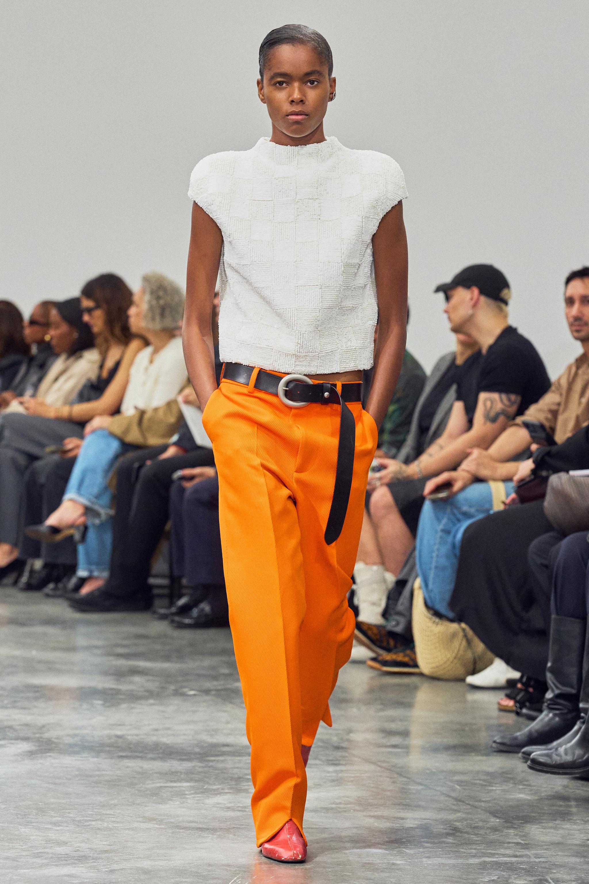 Fashion model walking runway wearing Courrèges modern white top and bright orange pants.