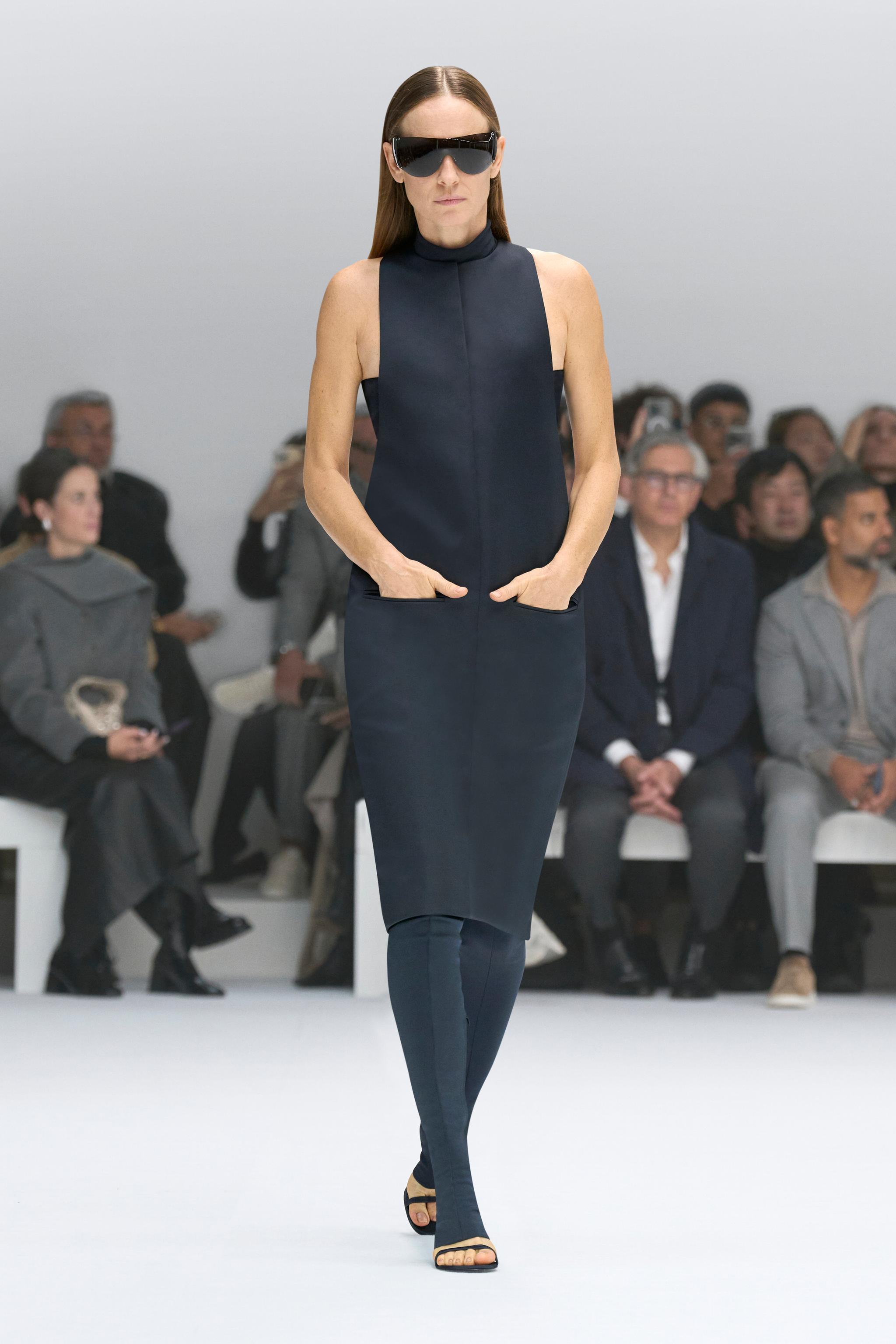 Elegant black sleeveless dress by Courrèges showcased on a runway with audience silhouettes.