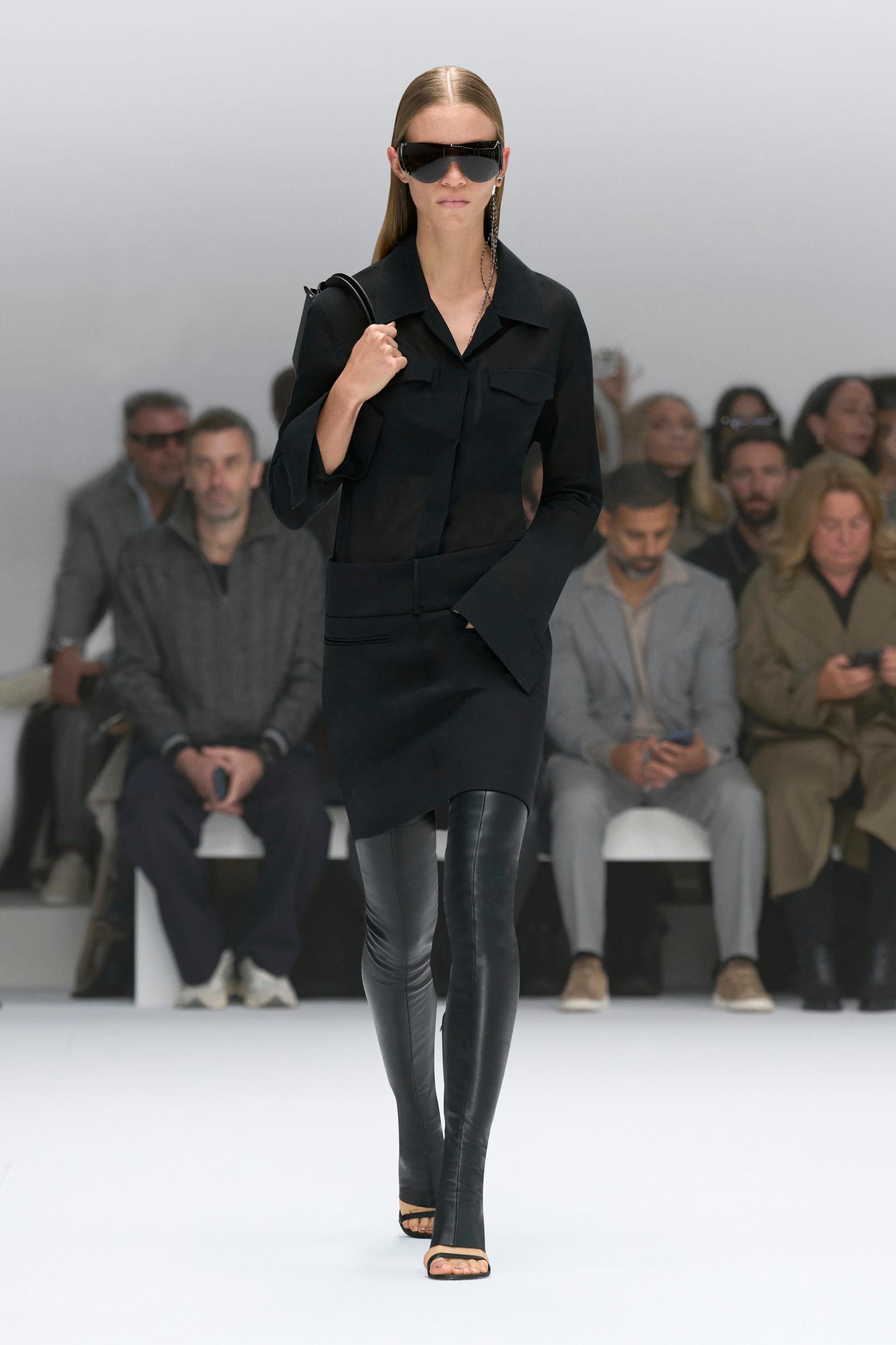 Elegant model in black Courrèges outfit walking on runway with audience in background.