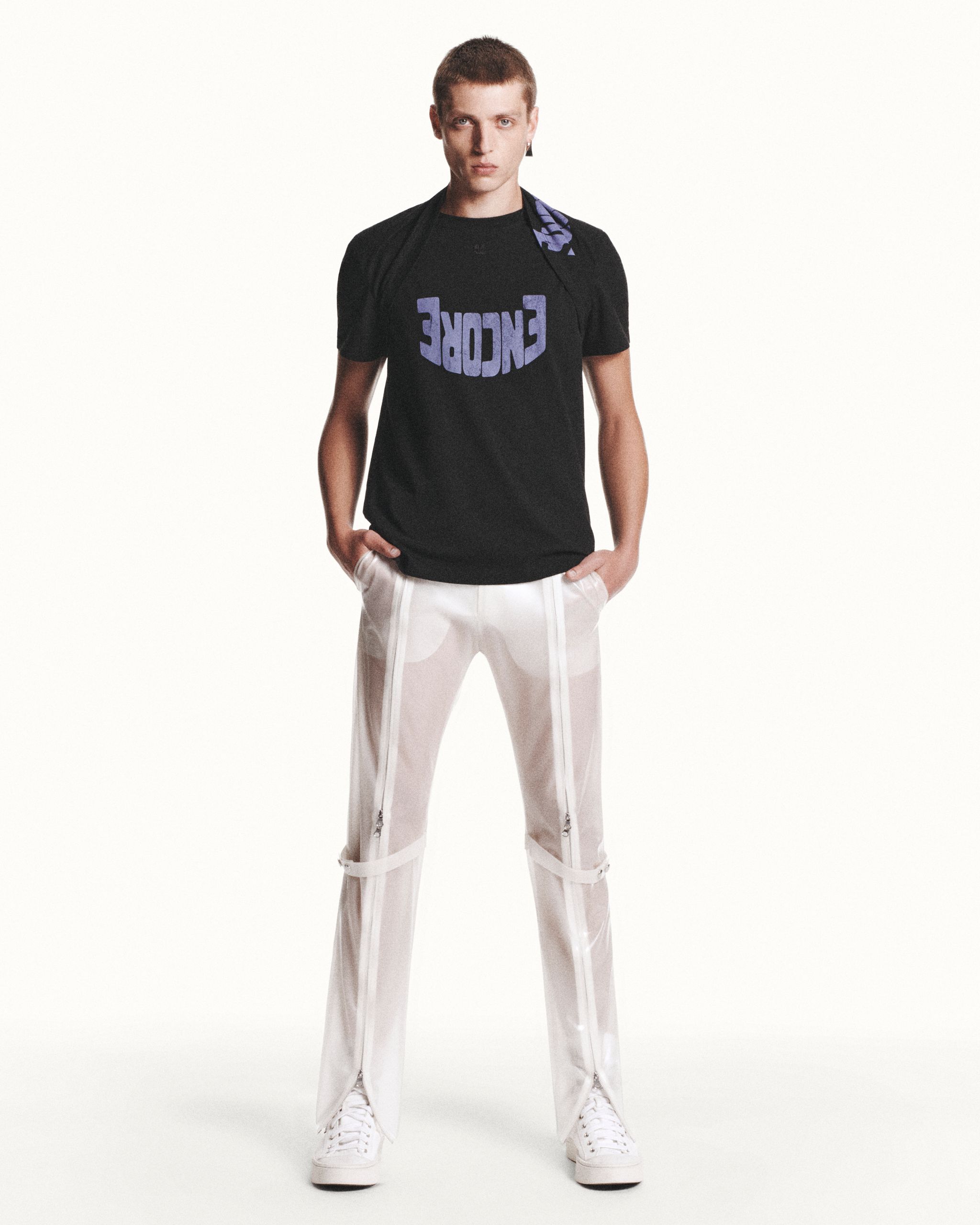 Young male model in black T-shirt and white pants, showcasing Courrèges fashion on a white background.