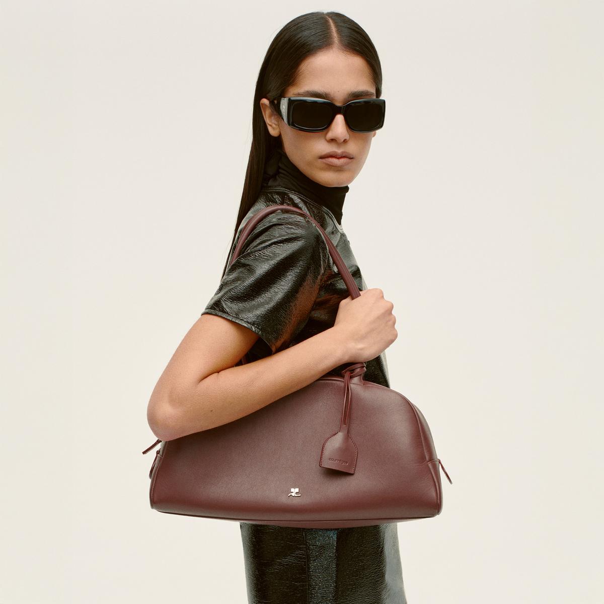 Elegant woman with dark sunglasses carrying a maroon designer handbag for Courrèges fashion.