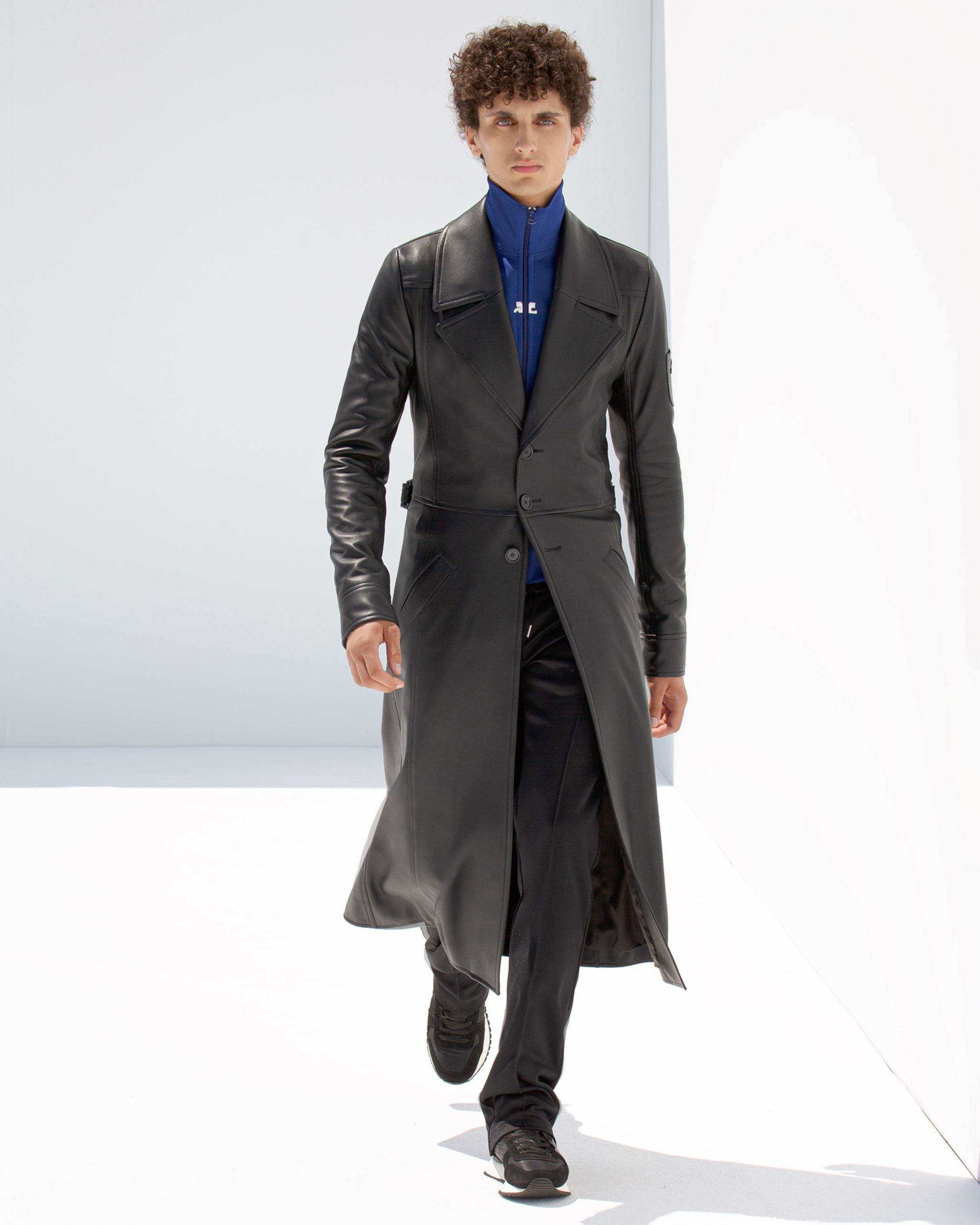 Elegant male model wearing a black leather coat and sporty zip-up blue top on a runway.