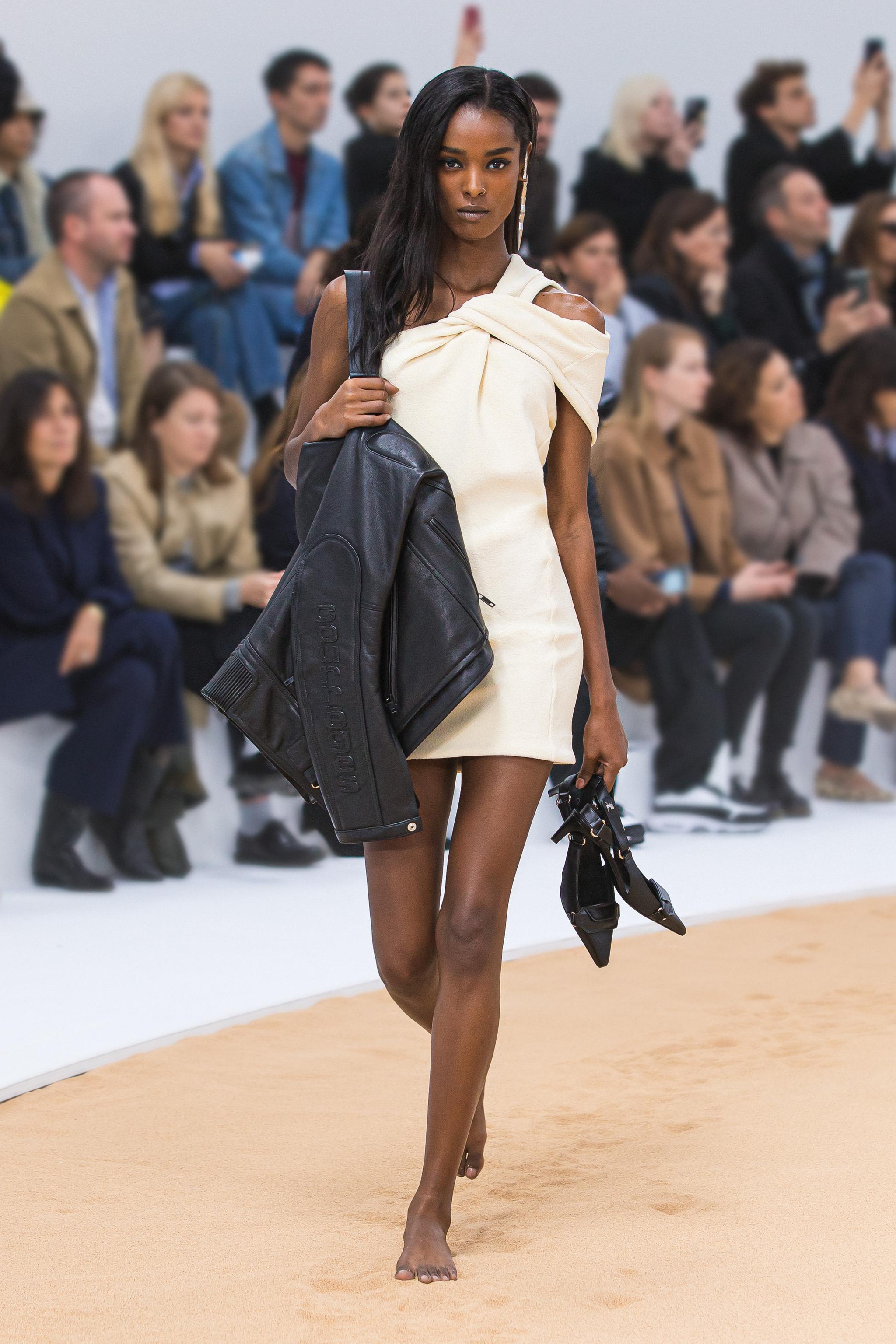 Modèle élégant en mode Courrèges sur runway, tenant une veste en cuir et des lunettes de soleil.
