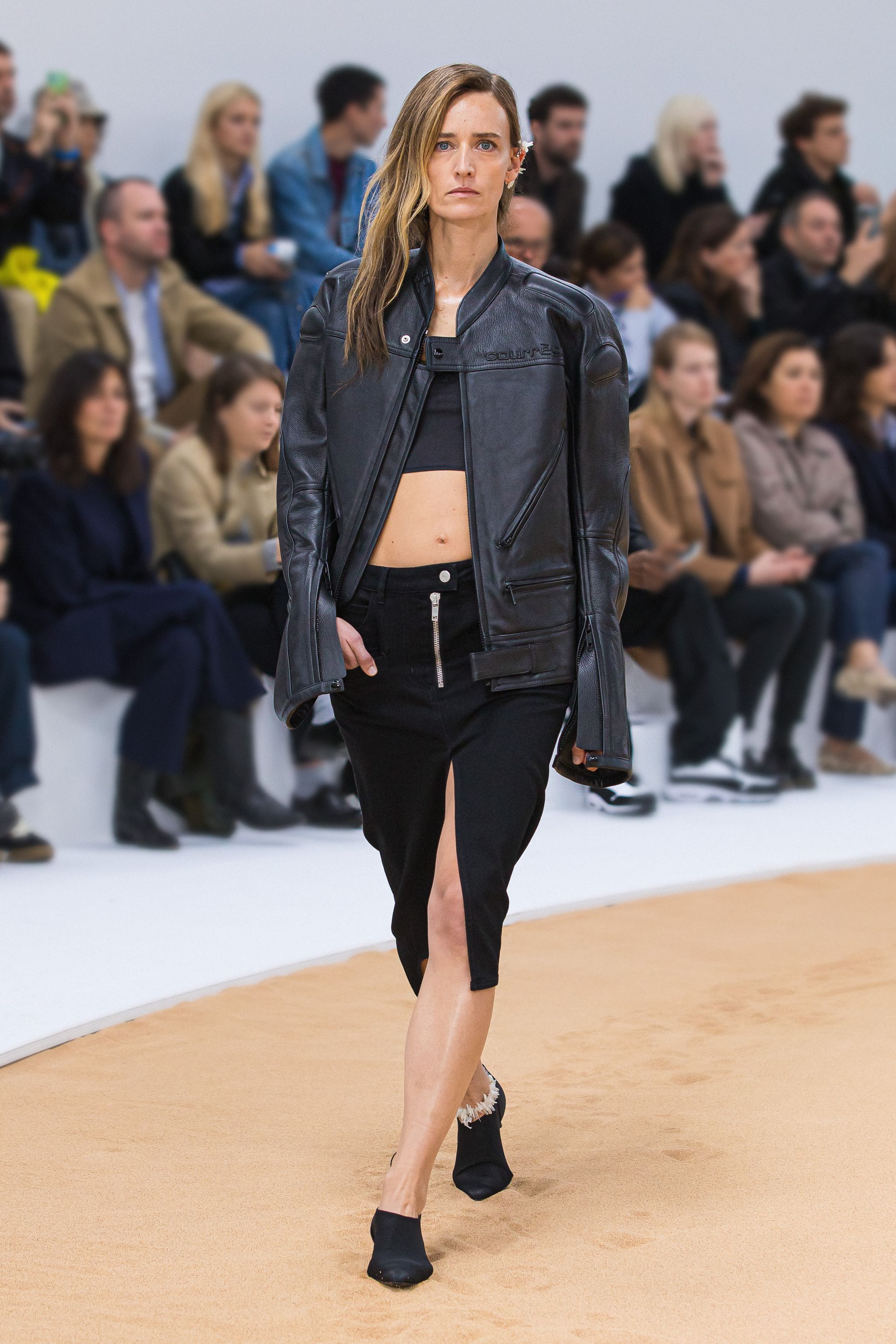 Fashion model walking runway in edgy black leather jacket and skirt showcasing Courrèges style.