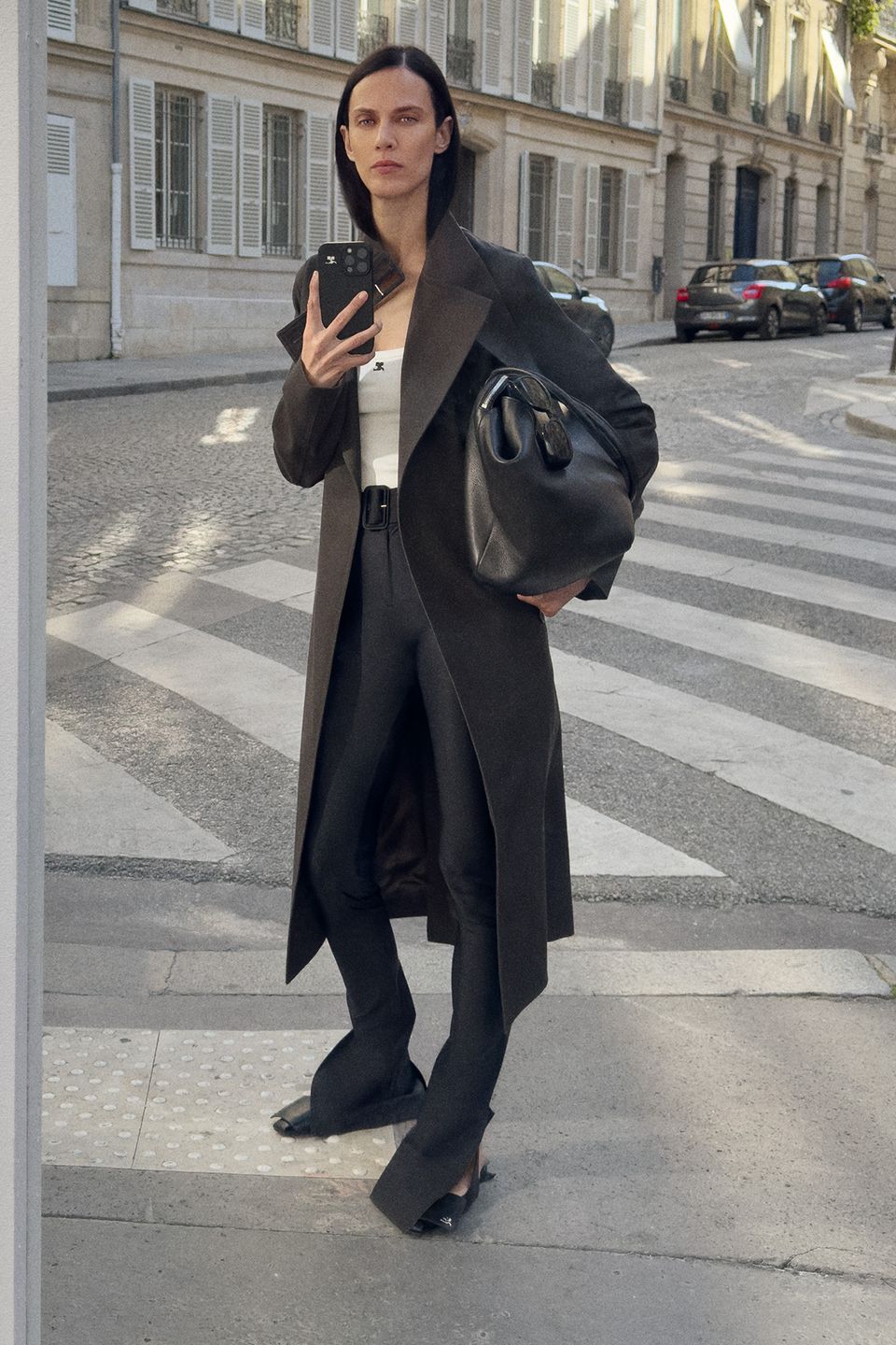 Chic woman in a trench coat and black trousers holding a designer handbag on a city street.