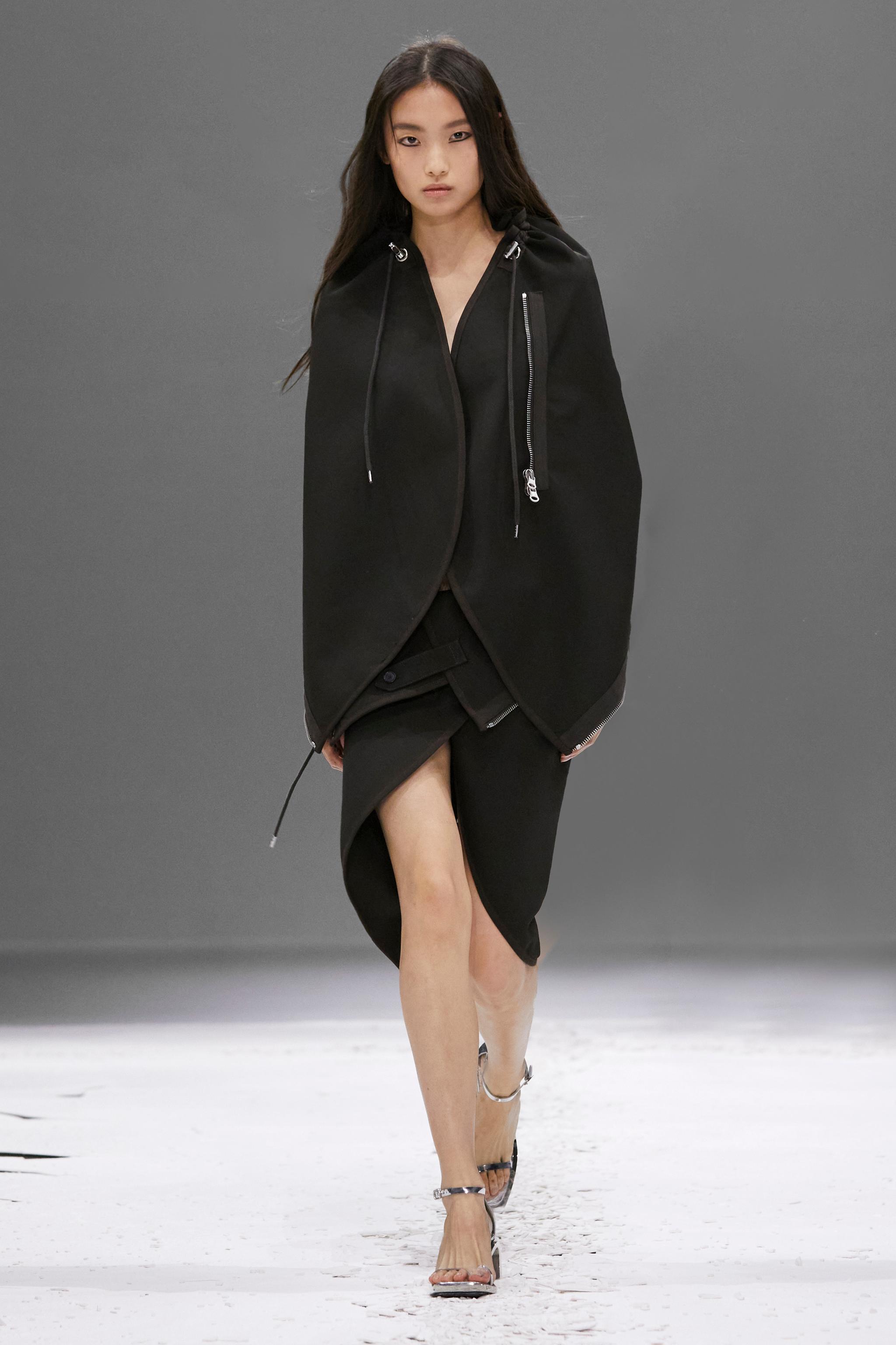 Elegant black Courrèges fashion model walking runway in a stylish hooded coat for an ecommerce site.