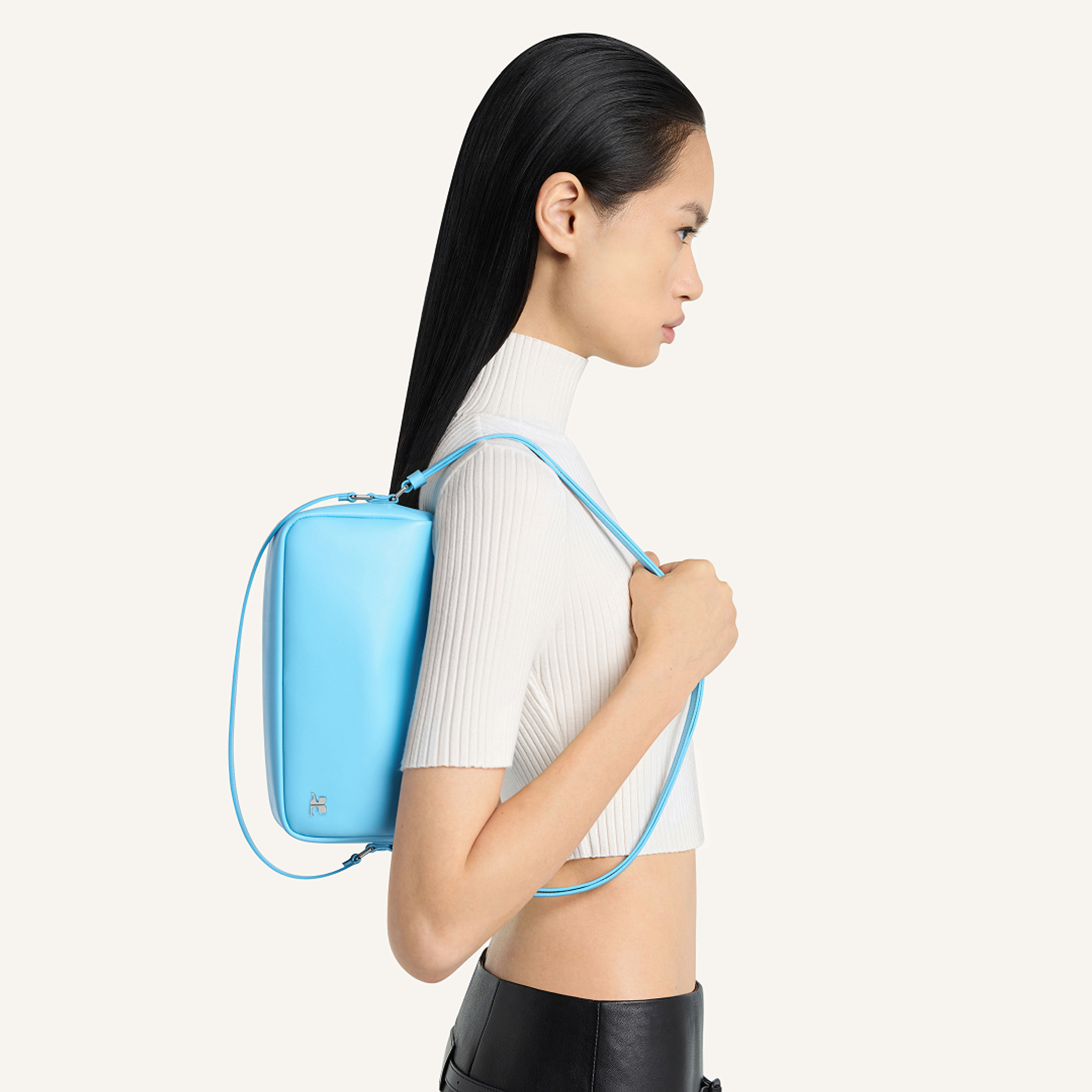 Elegant woman with sleek black hair wearing a white turtleneck, carrying a stylish blue Courrèges backpack.