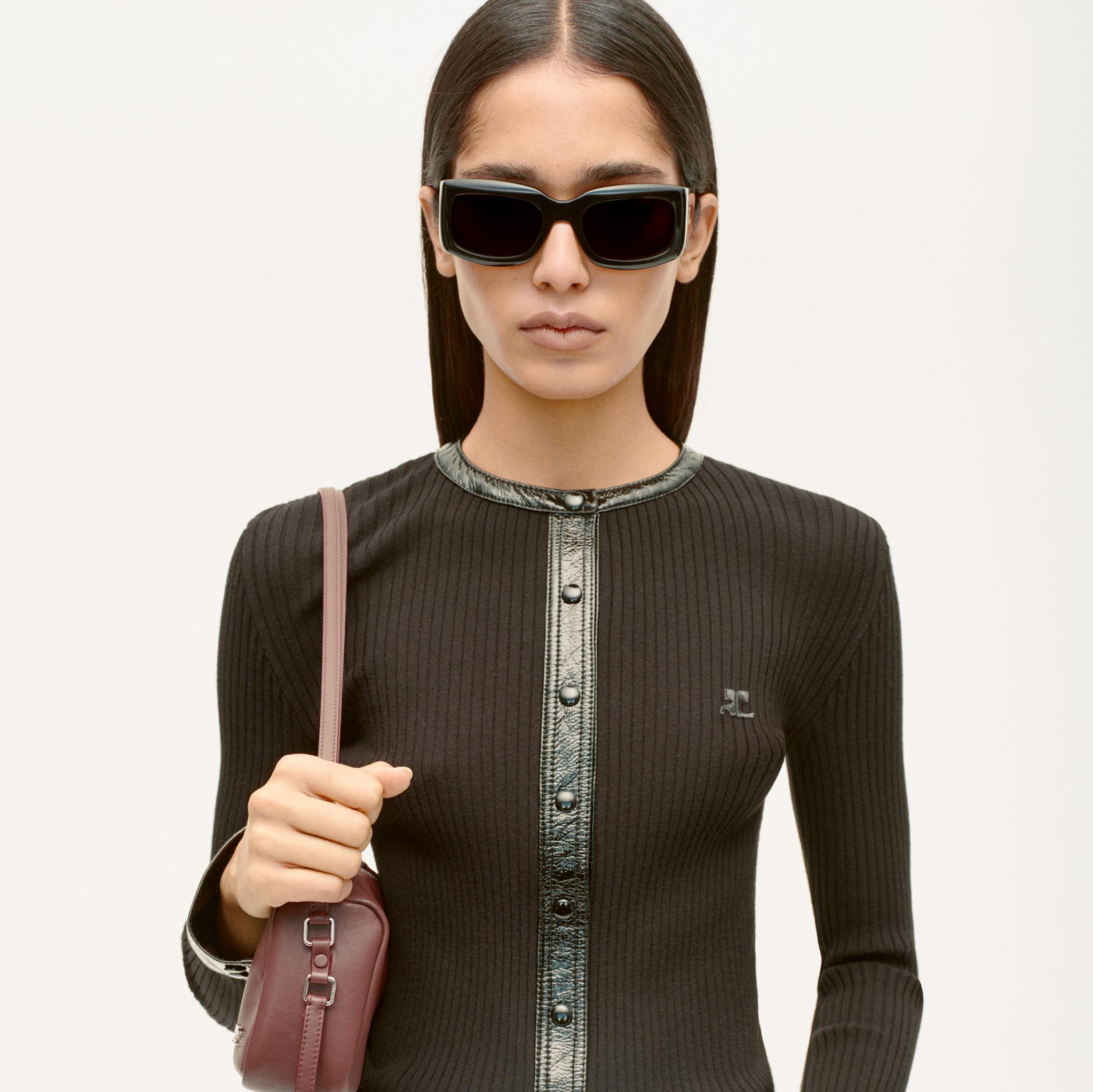 Femme élégante avec des lunettes de soleil noires, haut côtelé noir, et sac rose, représentant Courrèges.