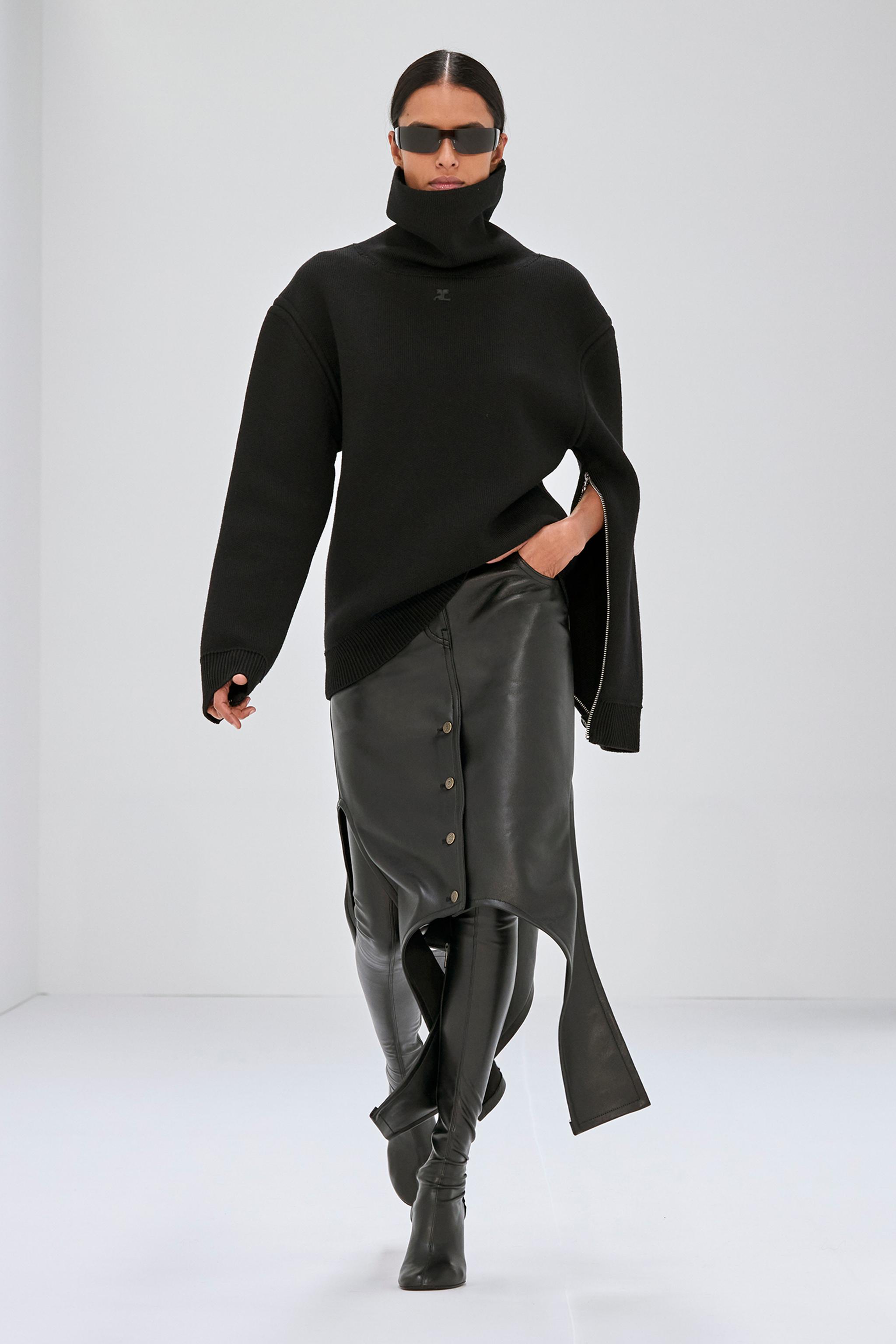 Pull noir élégant avec col roulé et coupe oversize, associé à une jupe en cuir et des bottes, par Courrèges.