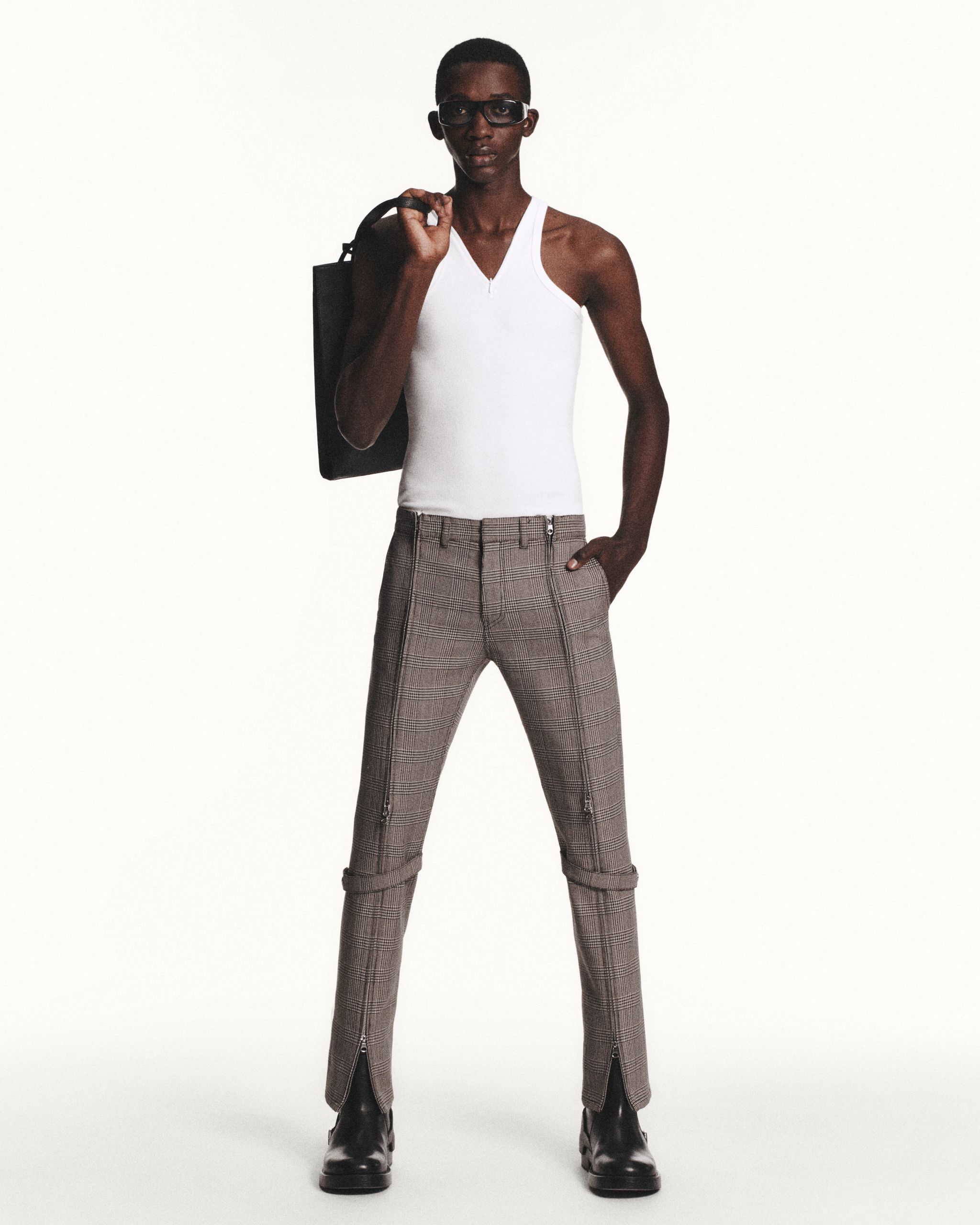 Elegant male model wearing Courrèges plaid pants, white tank top, with black boots, accessory, and glasses.