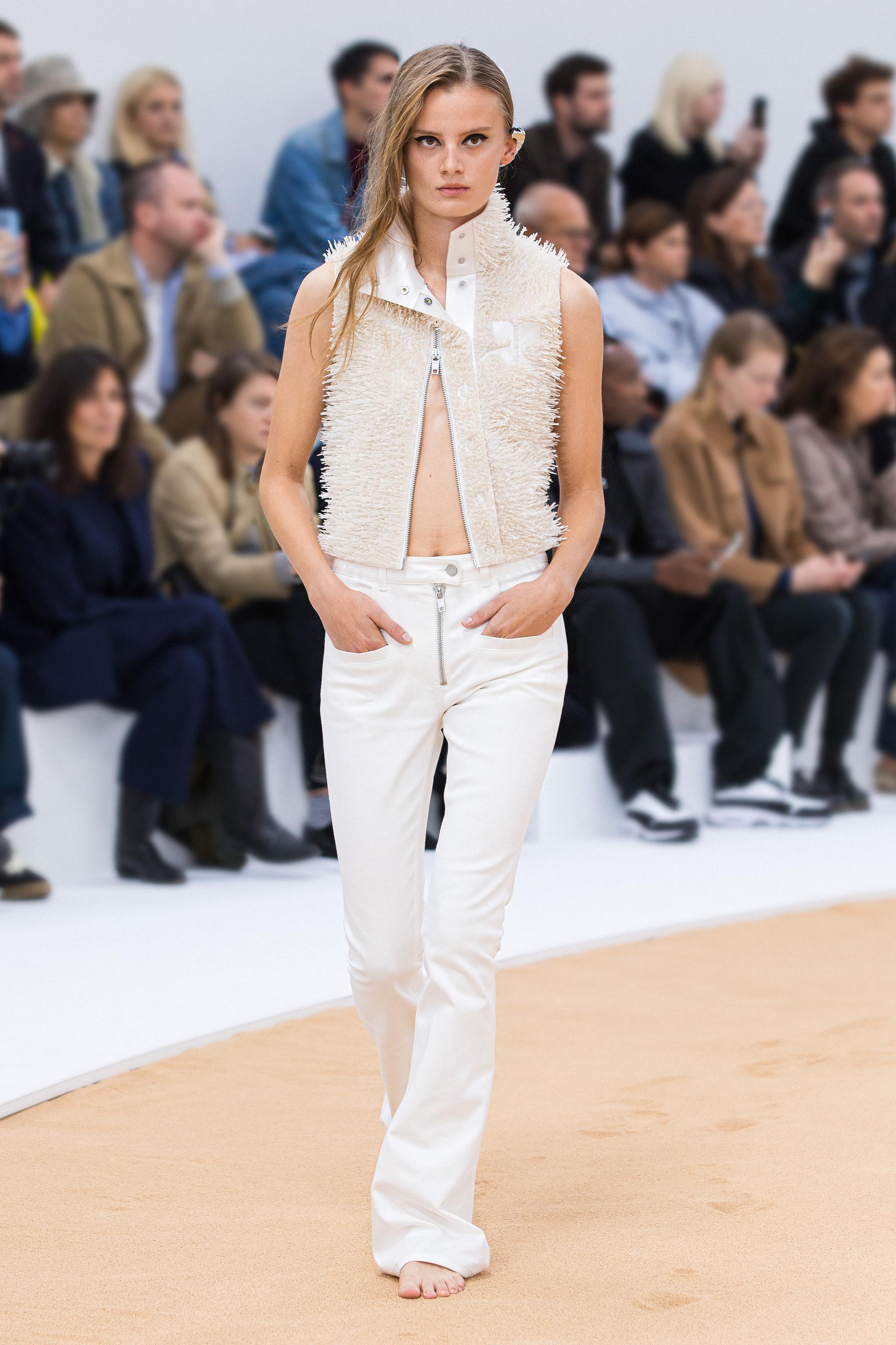 Modèle femme élégante défilant en Courrèges avec un gilet crème et pantalon blanc, lors d'un défilé.