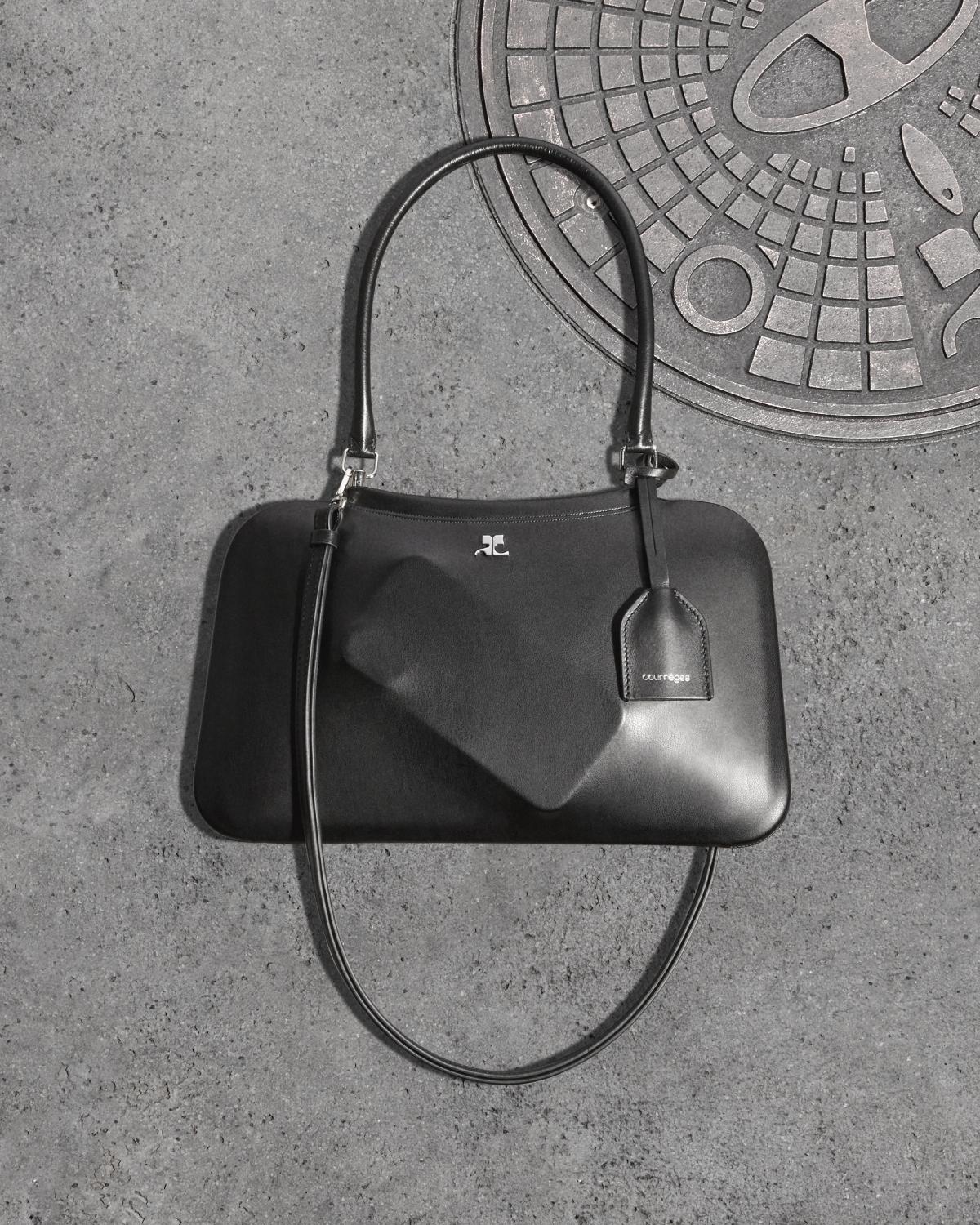 Elegant black Courrèges handbag with a sleek design, placed on a concrete surface near a manhole cover.
