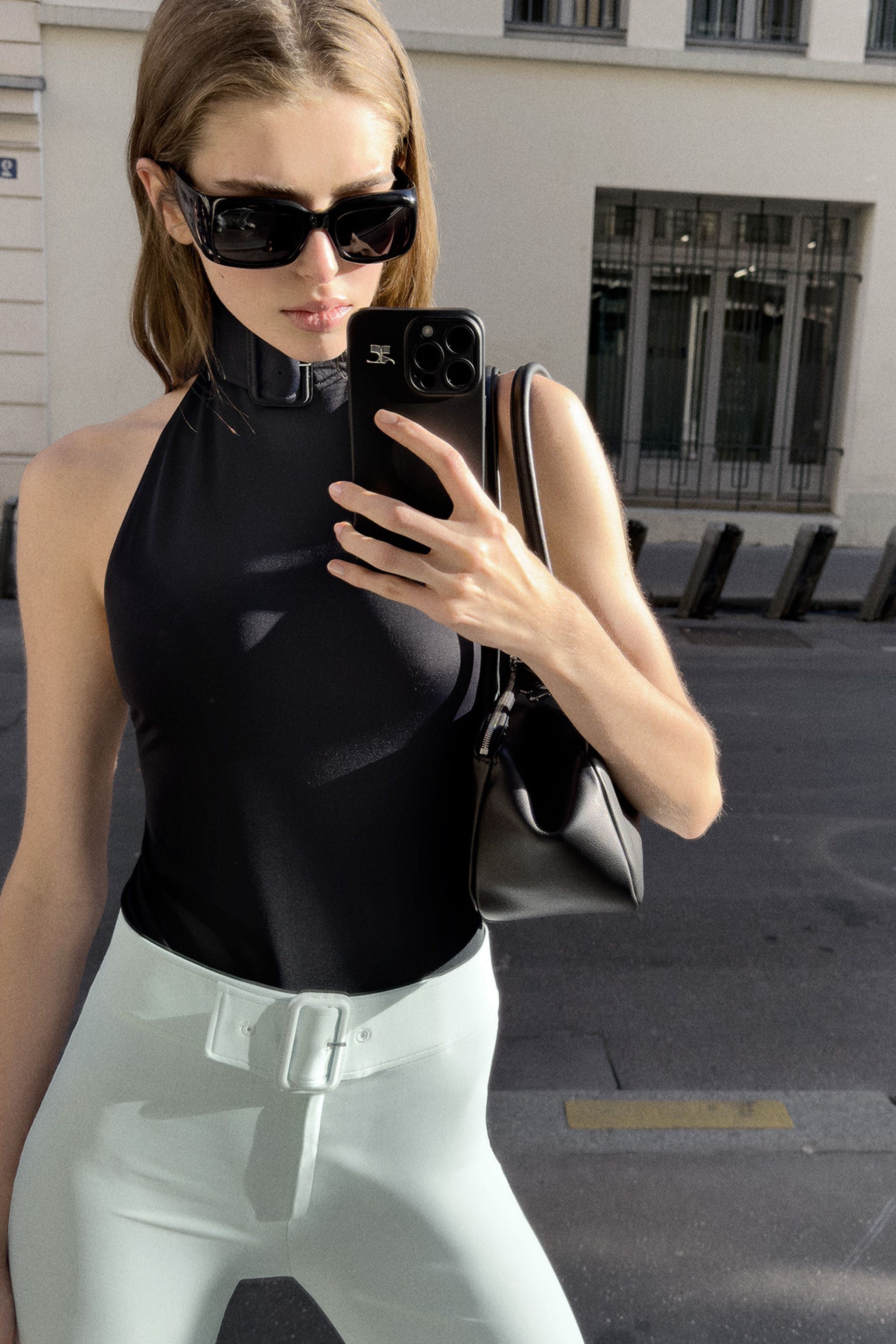 Elegant woman in black top and white pants take a mirror selfie on an urban street for Courrèges.