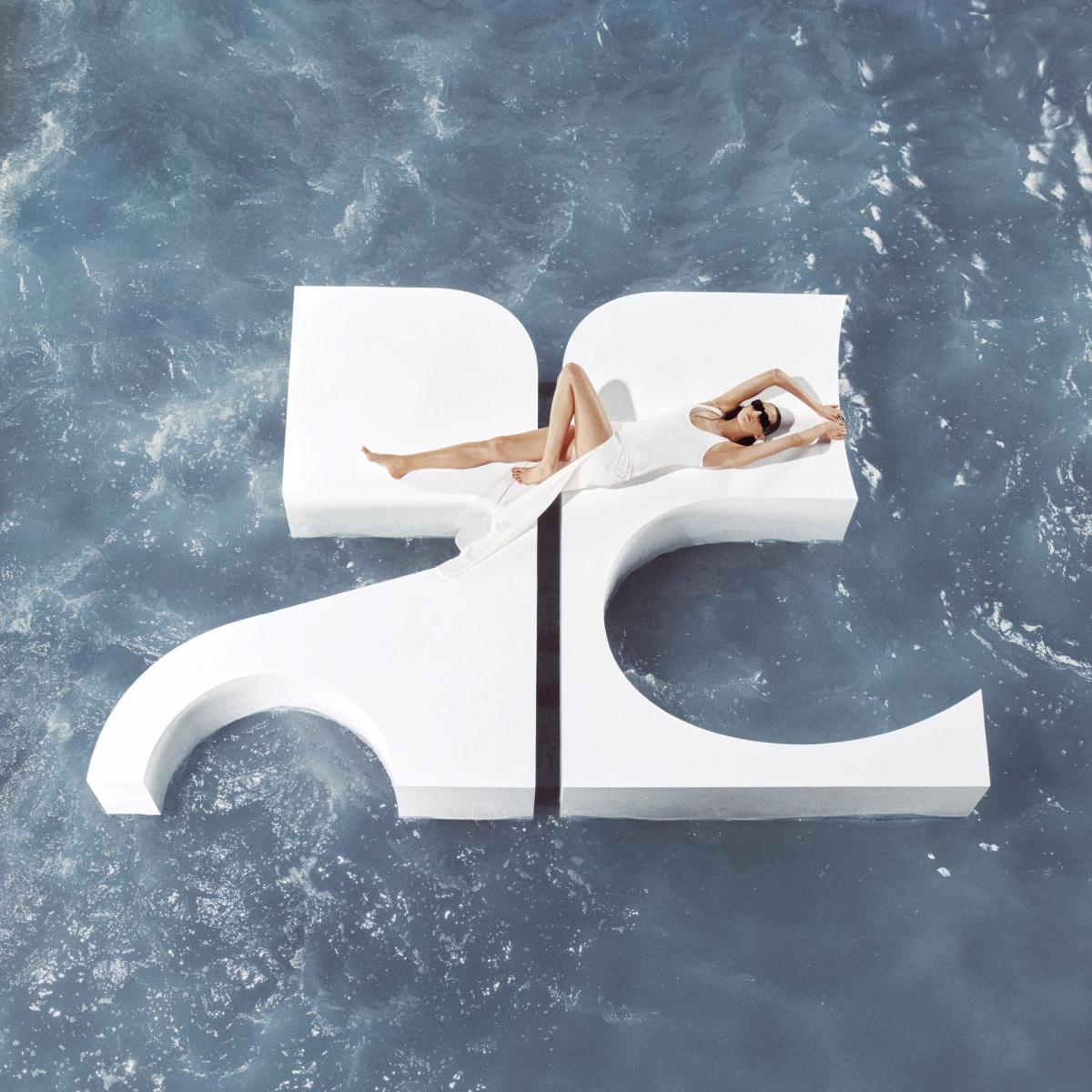 Elegant model in white fashion attire lounging on a giant Courrèges logo near water.