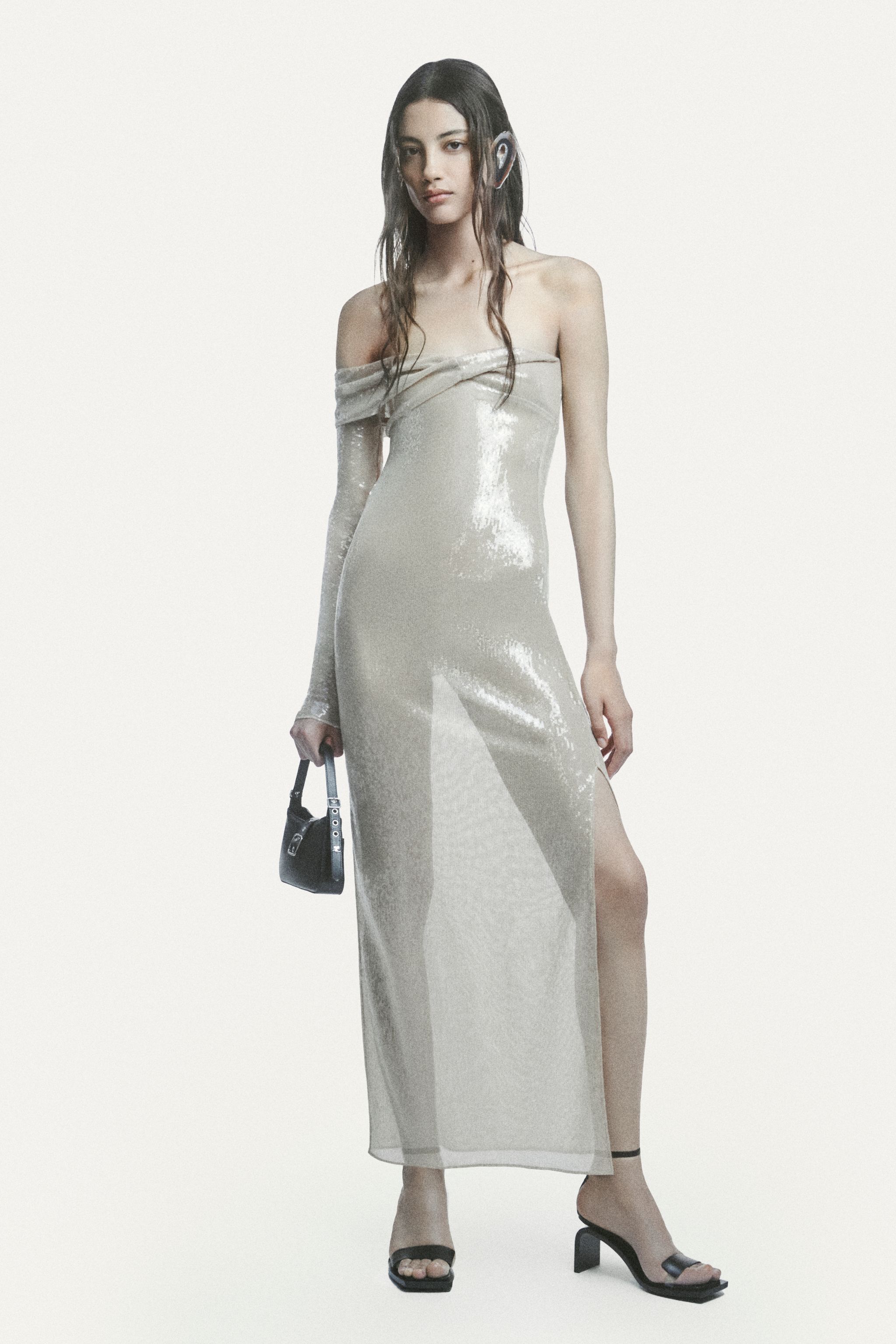 Elegant model in shiny off-shoulder dress holding a small black handbag, high heels, on a white background.