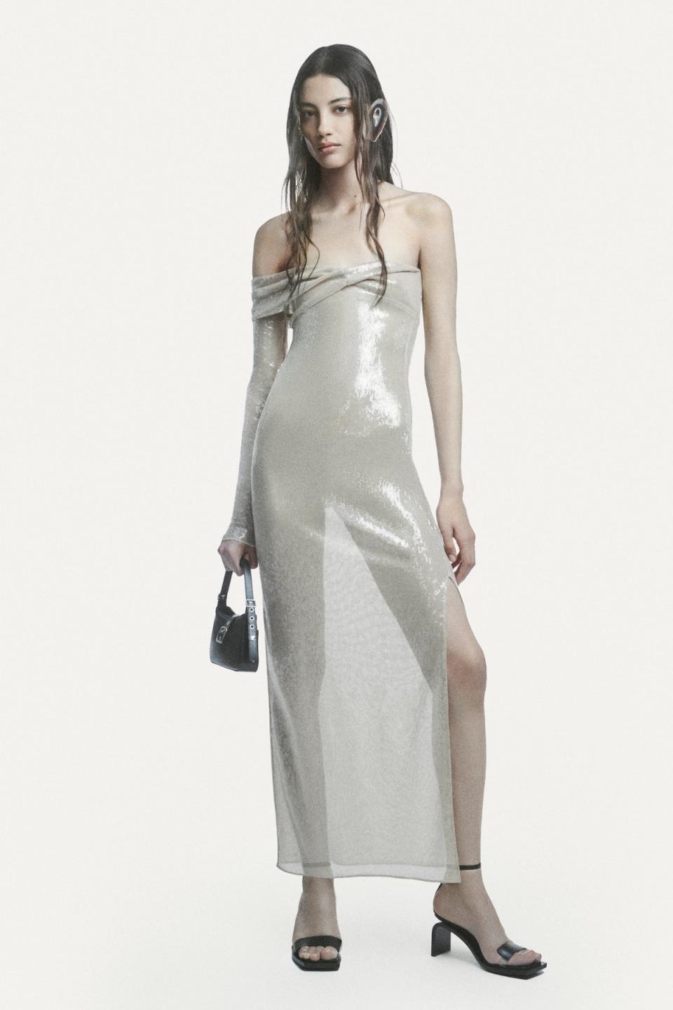 Elegant model in shiny off-shoulder dress holding a small black handbag, high heels, on a white background.