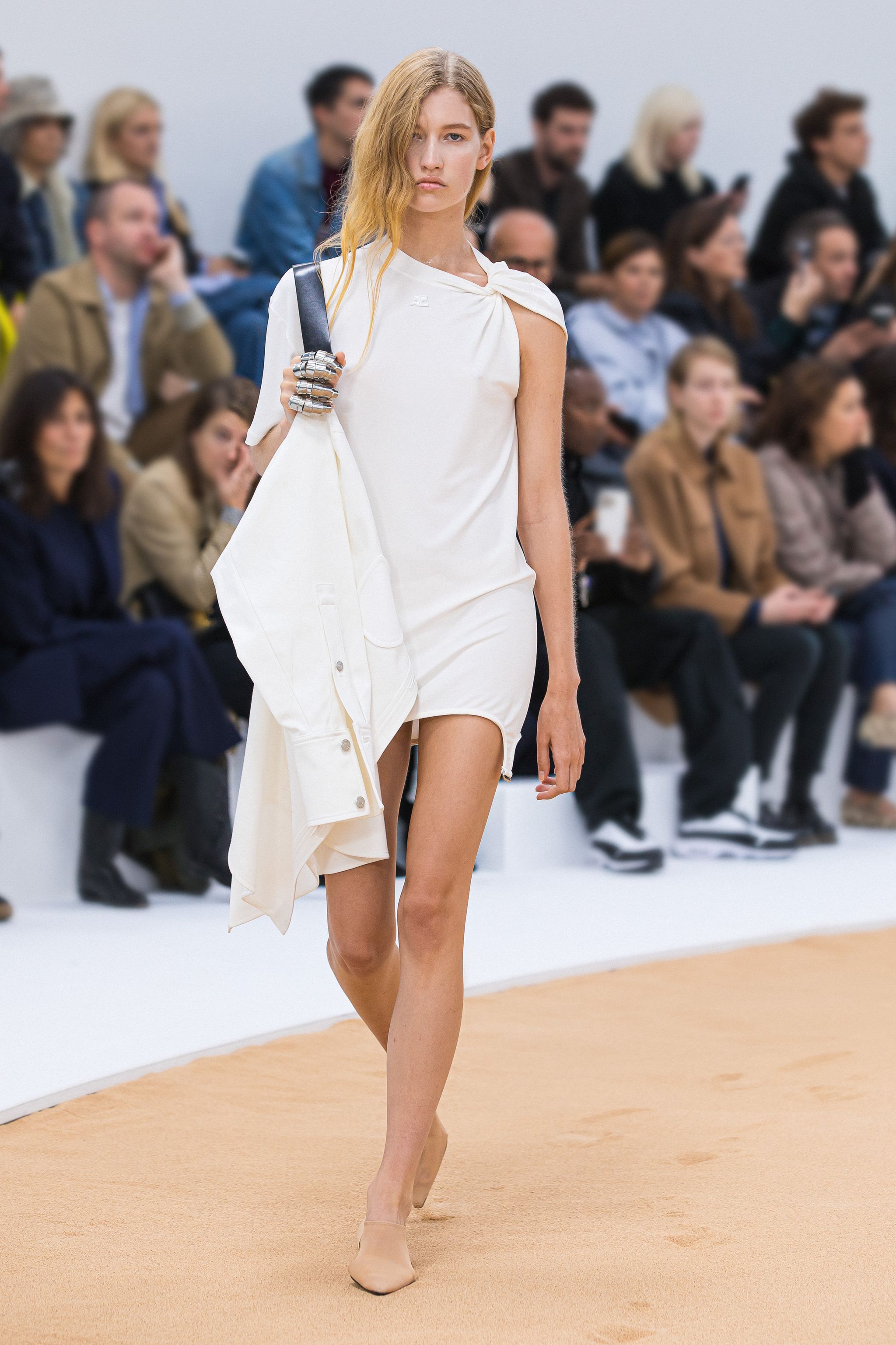 Elegant white dress model walking on runway at Courrèges fashion show, surrounded by audience.