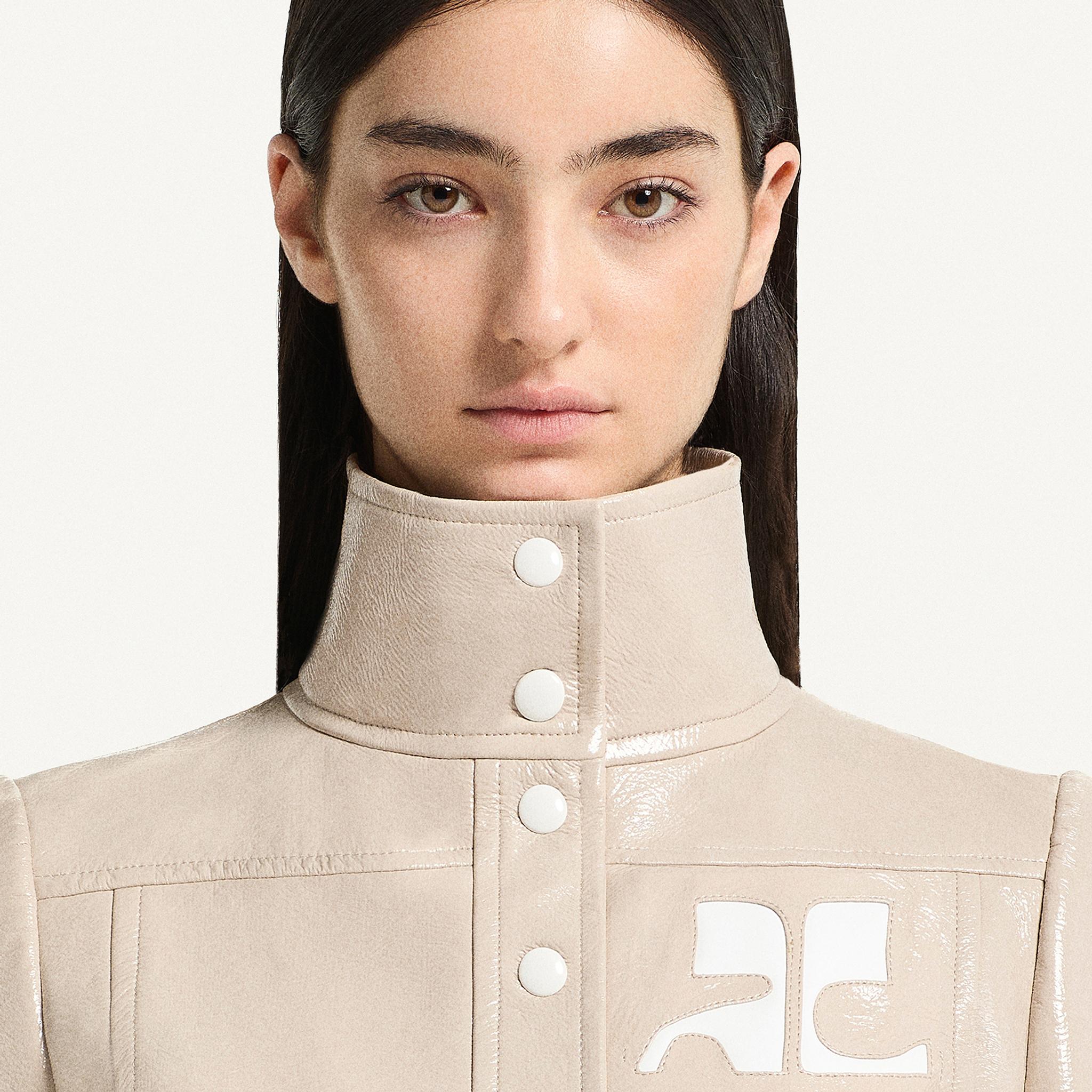 Elegant beige Courrèges high-neck jacket with white logo and snap buttons, on a model.