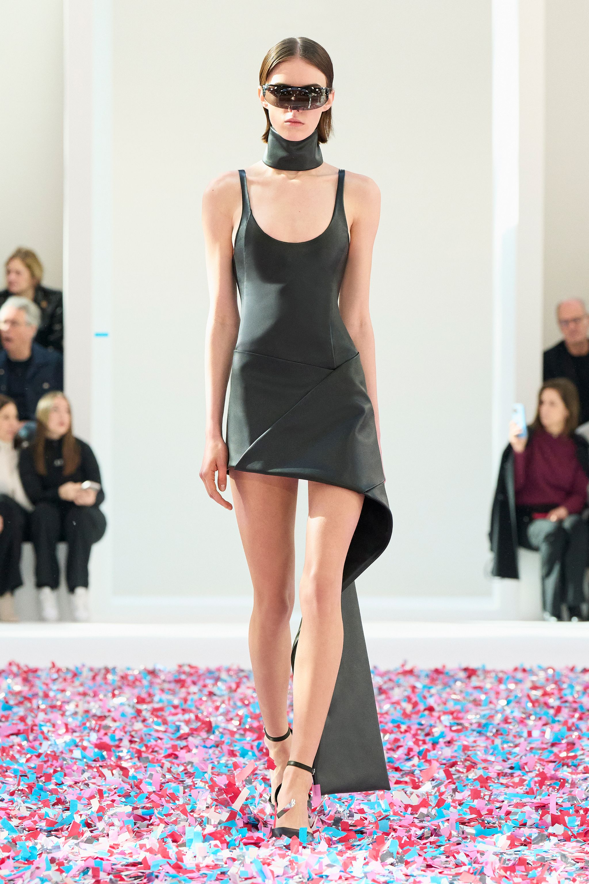 Fashion model walking on a runway in a sleek black dress with futuristic sunglasses for Courrèges.